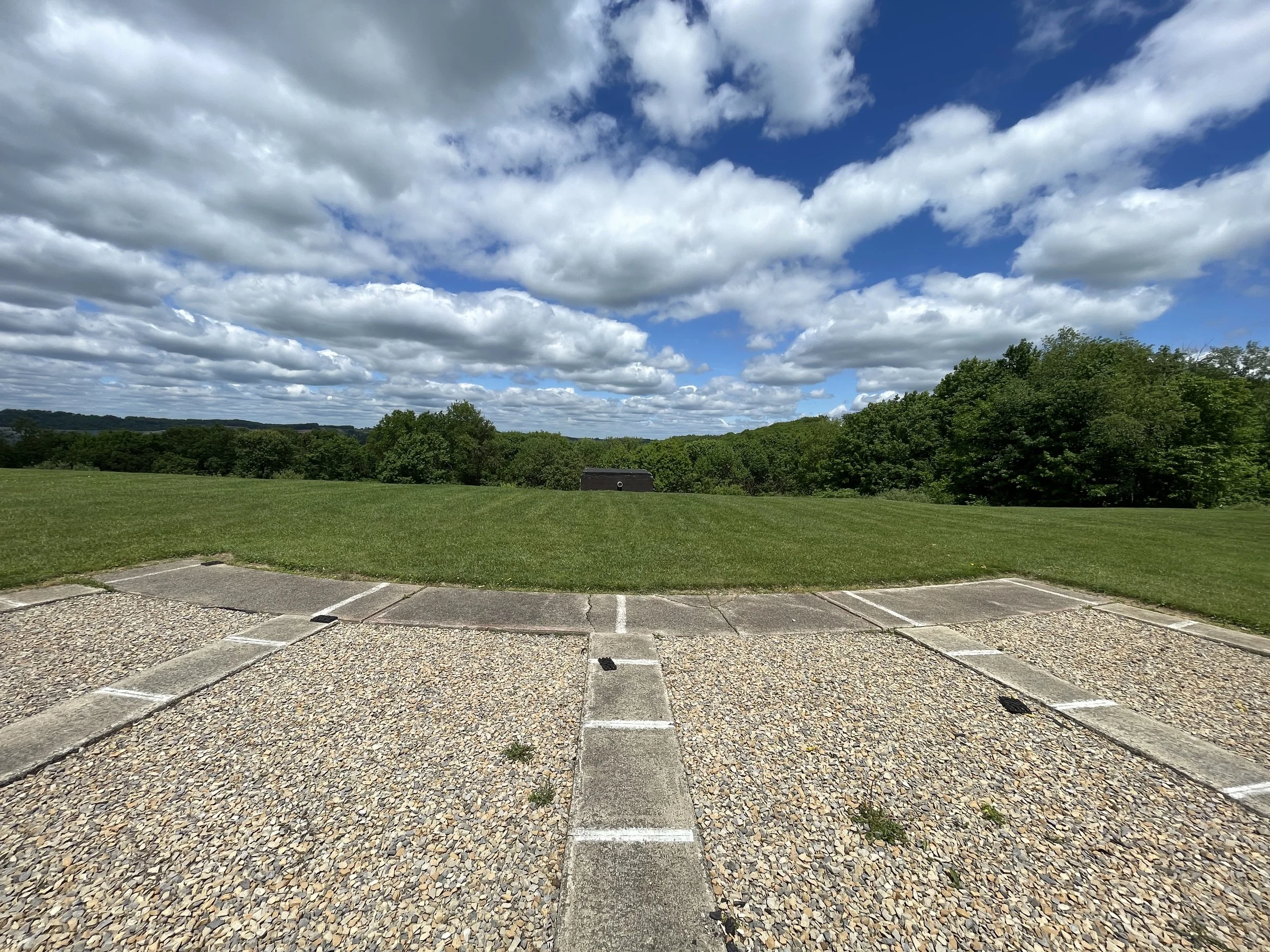 Empty parking lot with gravel and concrete spaces, grassy field, trees, blue sky with scattered clouds in a rural setting.