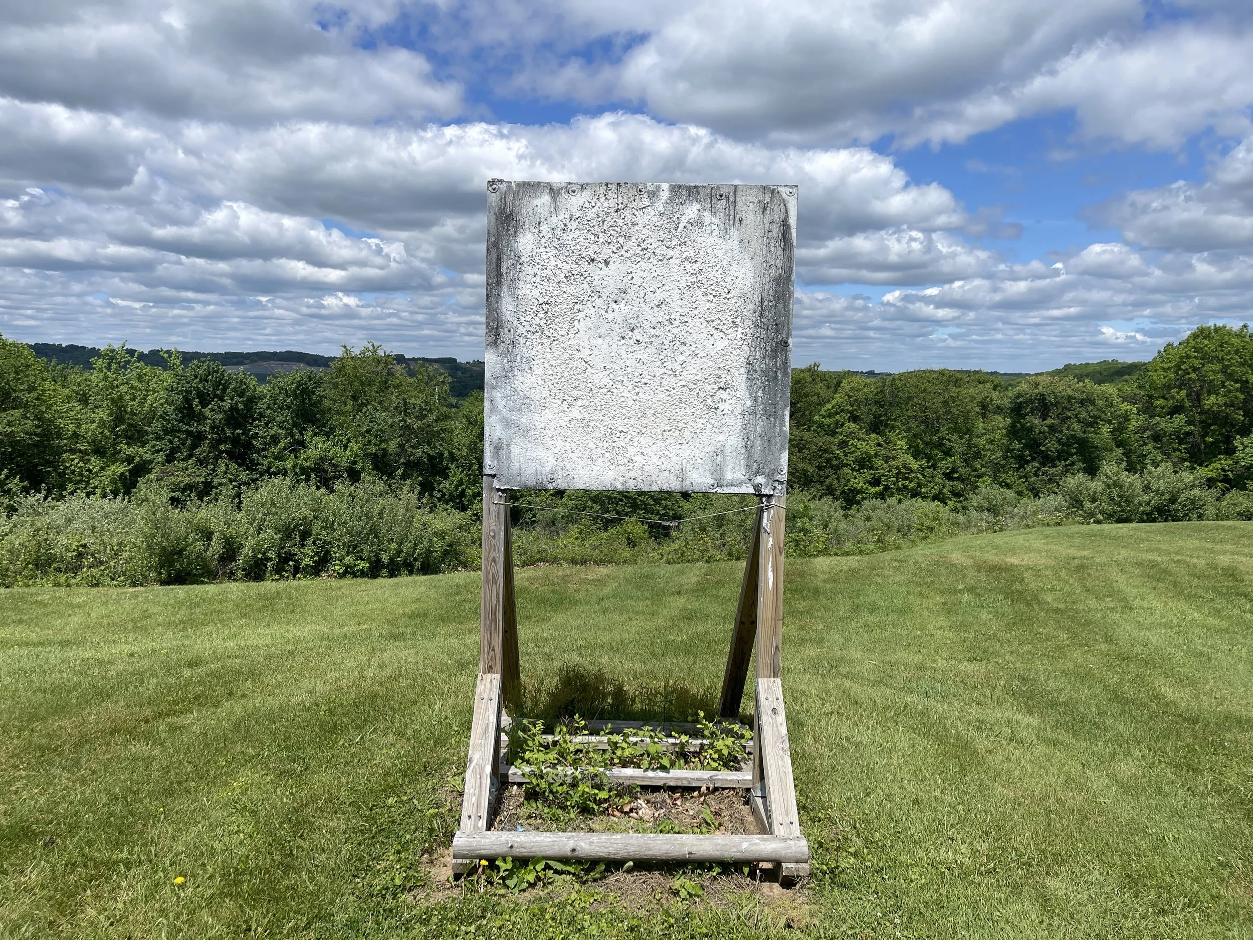 Empty weathered board on wooden stand outdoors with green grass, trees, and cloudy sky in the background.