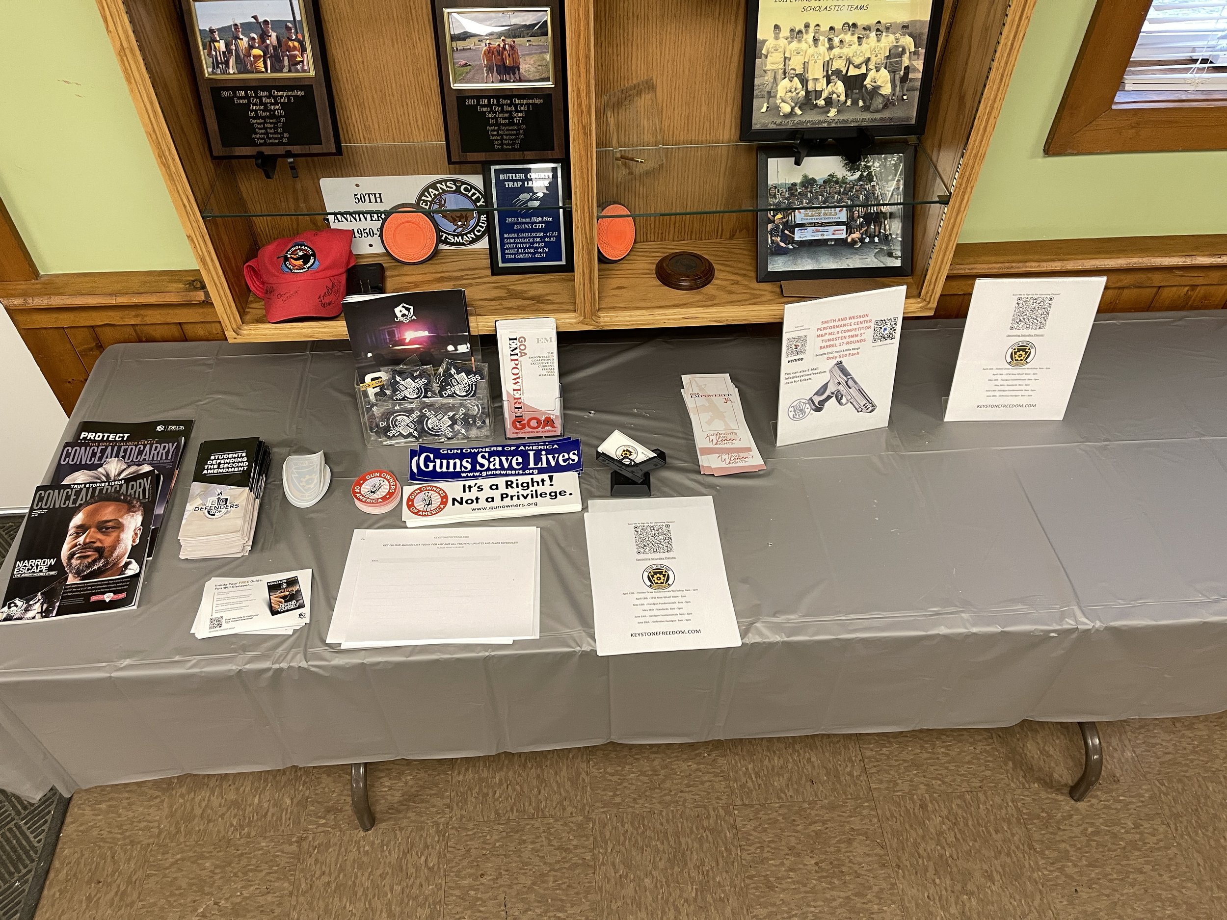 Table with various pamphlets, stickers, and flyers about gun rights and legislation, with framed photos of sports teams and awards in the background.
