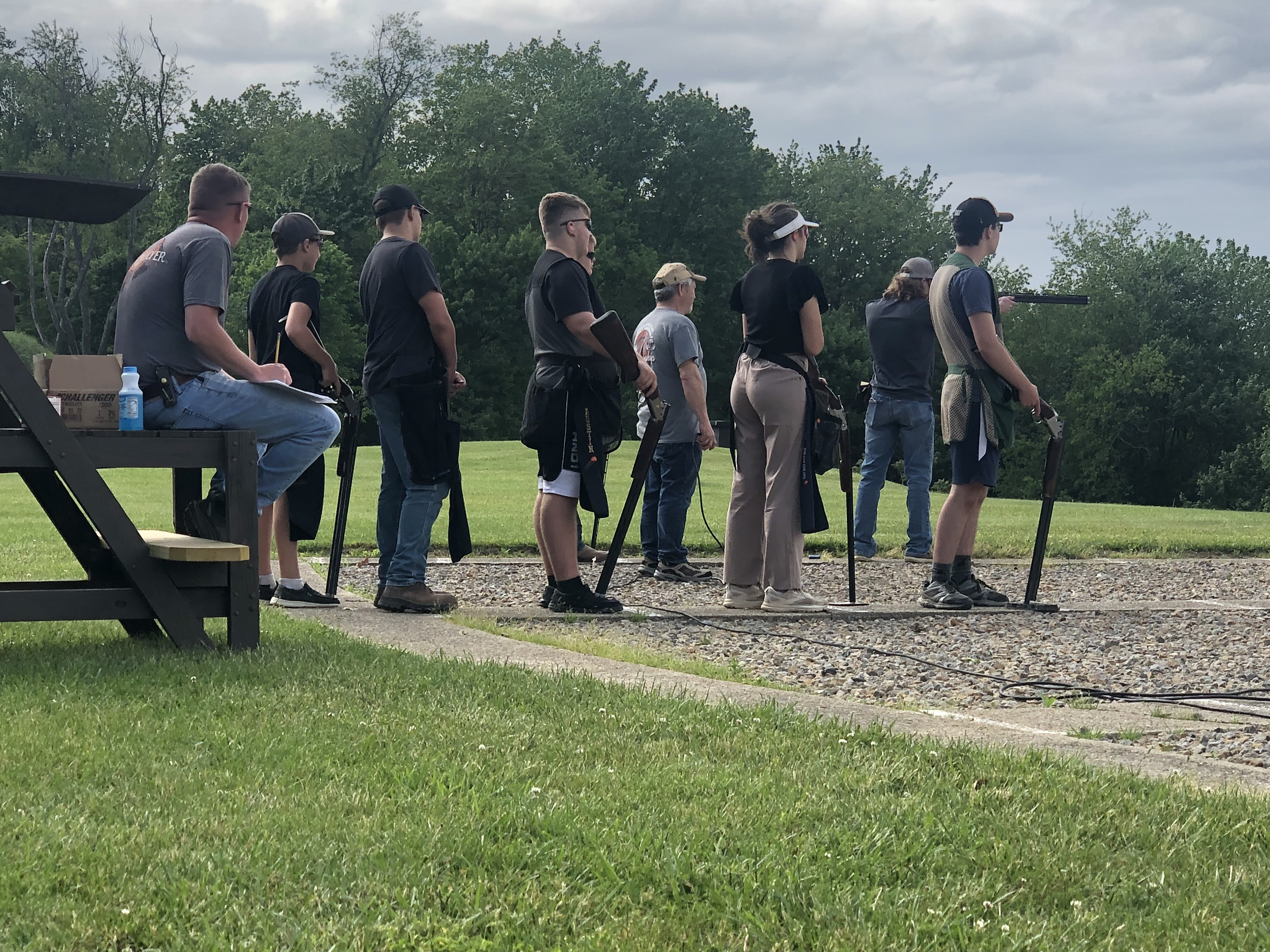 A group of people practicing shooting clay pigeons at an outdoor range, standing in a line with shotguns, with one person seated nearby.
