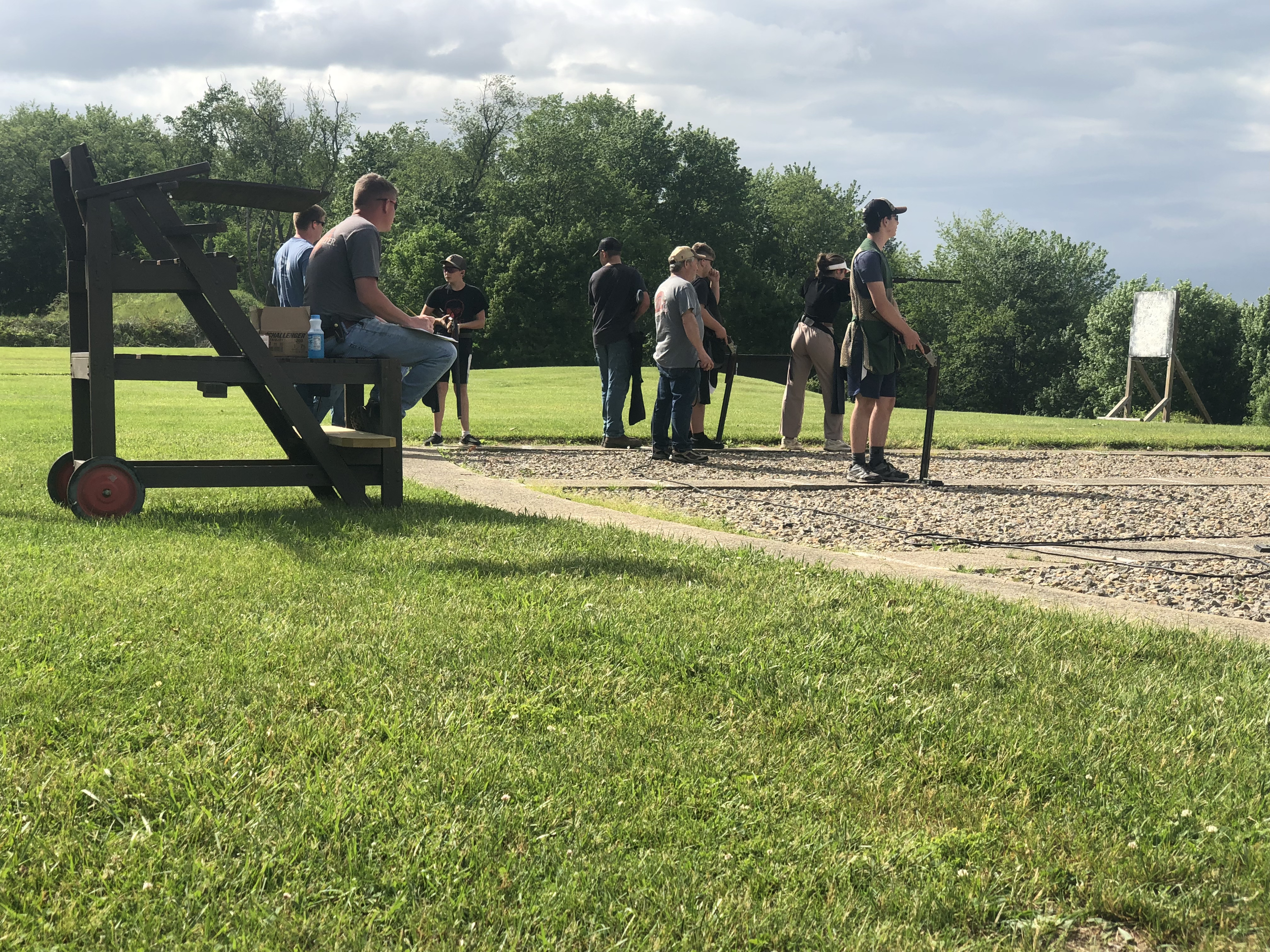 Group of people participating in a shooting activity outdoors on a grassy field, with some aiming rifles and others observing.