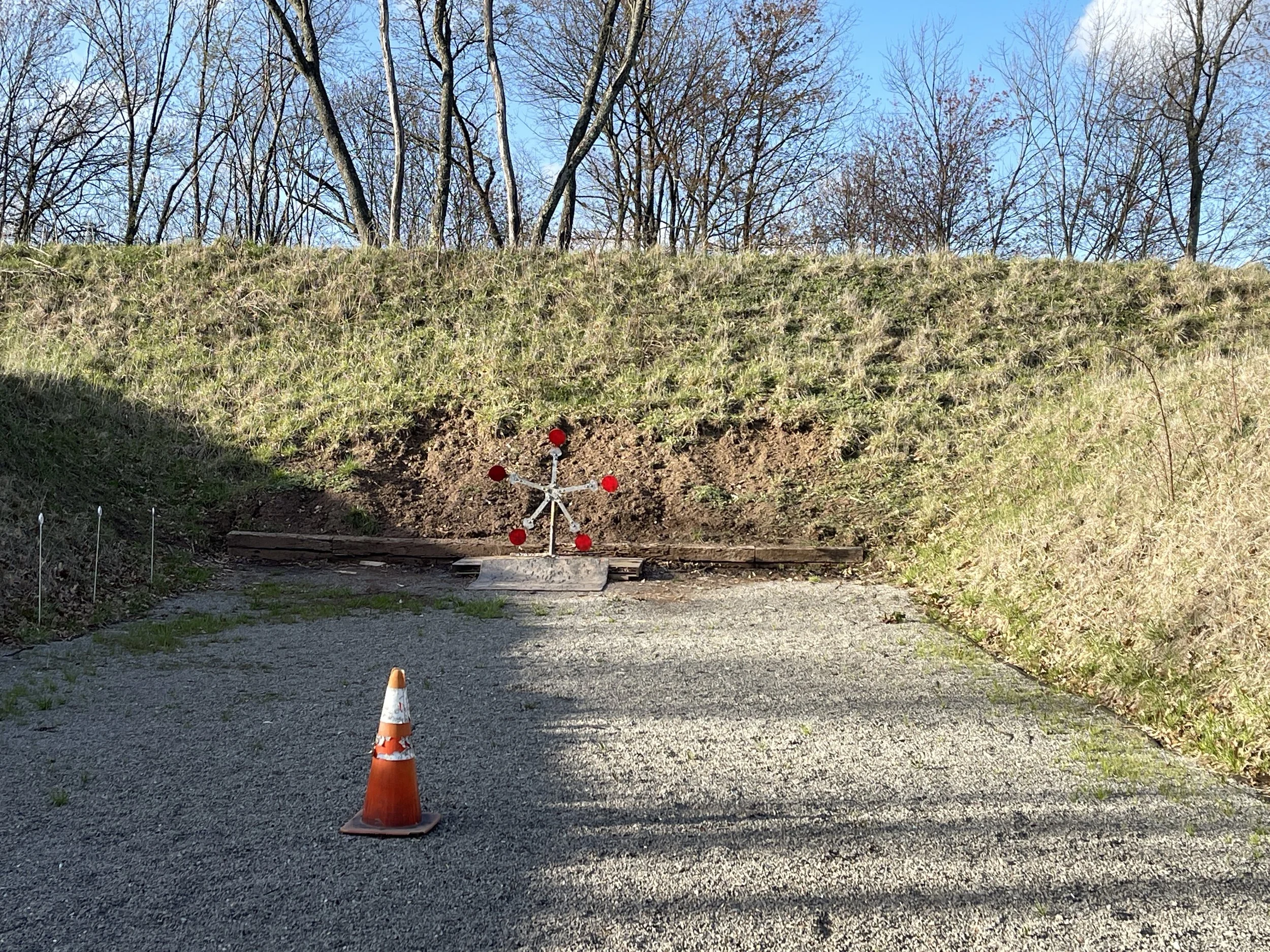 A small target practice stand with red dots on it, placed on a gravel area in front of a grassy embankment, with an orange traffic cone nearby, and trees in the background.