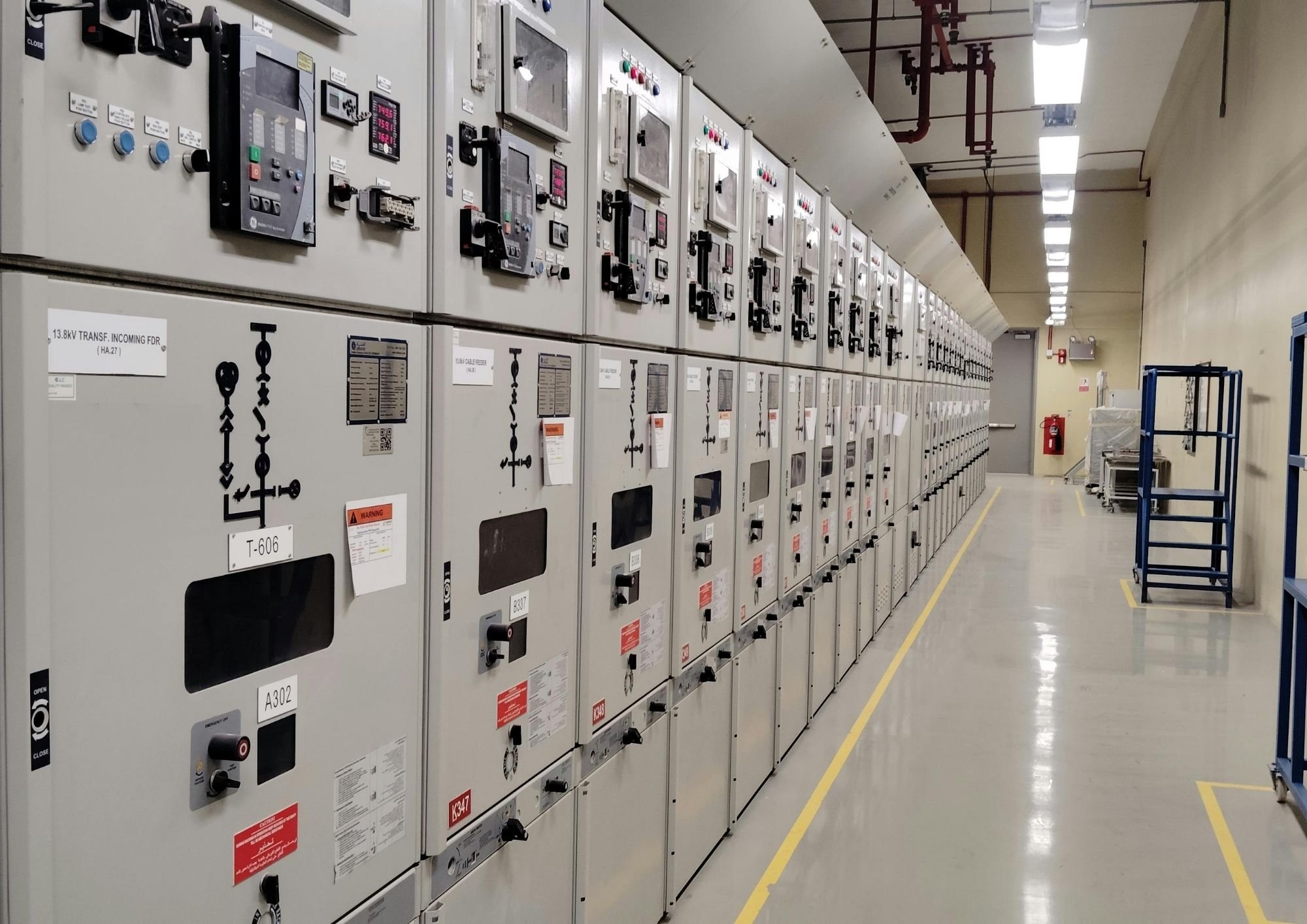 An aisle in an industrial or data facility with electrical control panels lining the wall, red pipes overhead, and a blue cart on the right side.