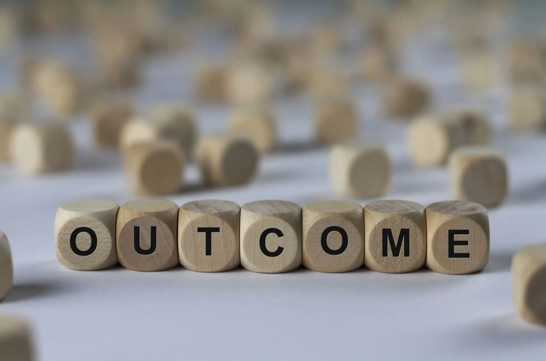 Wooden blocks spelling out the word 'OUTCOME' with blurred additional blocks and scattered pieces in the background.