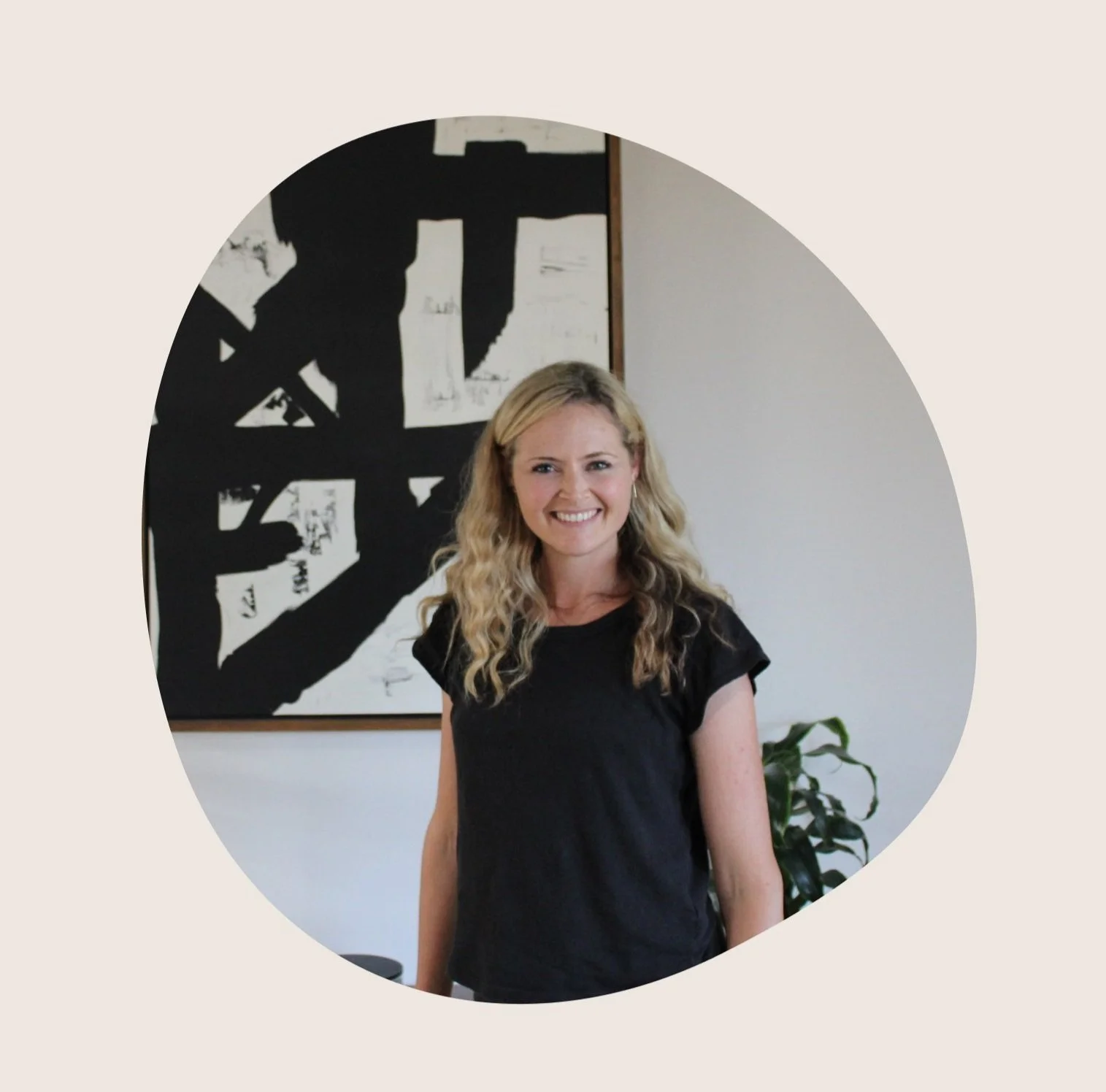 A smiling woman with long blonde curly hair wearing a black t-shirt, standing in front of an abstract black and white artwork and a green plant in an office or gallery setting.