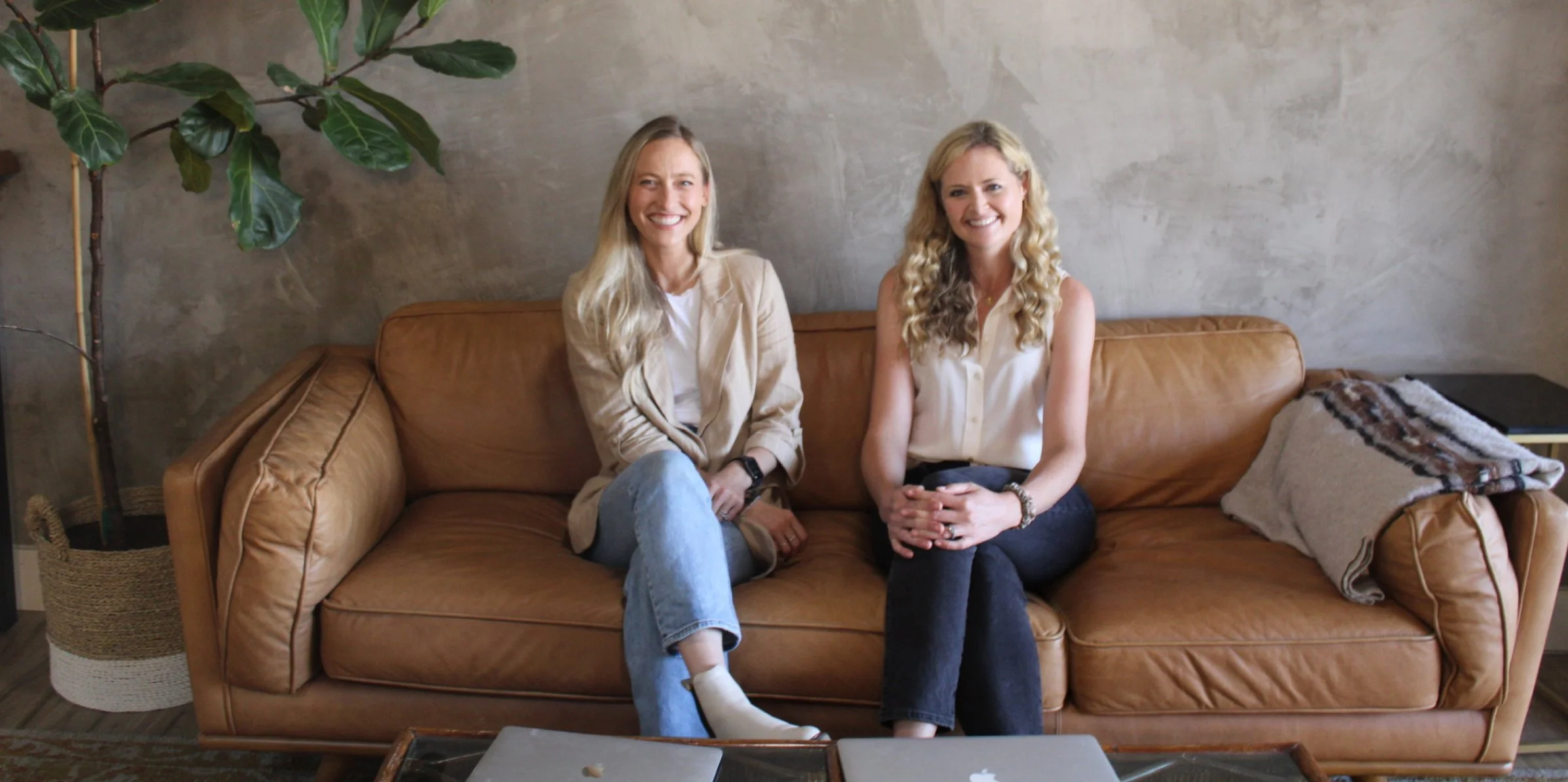 Two women sitting on a brown leather couch in a modern living room, smiling and looking at the camera.