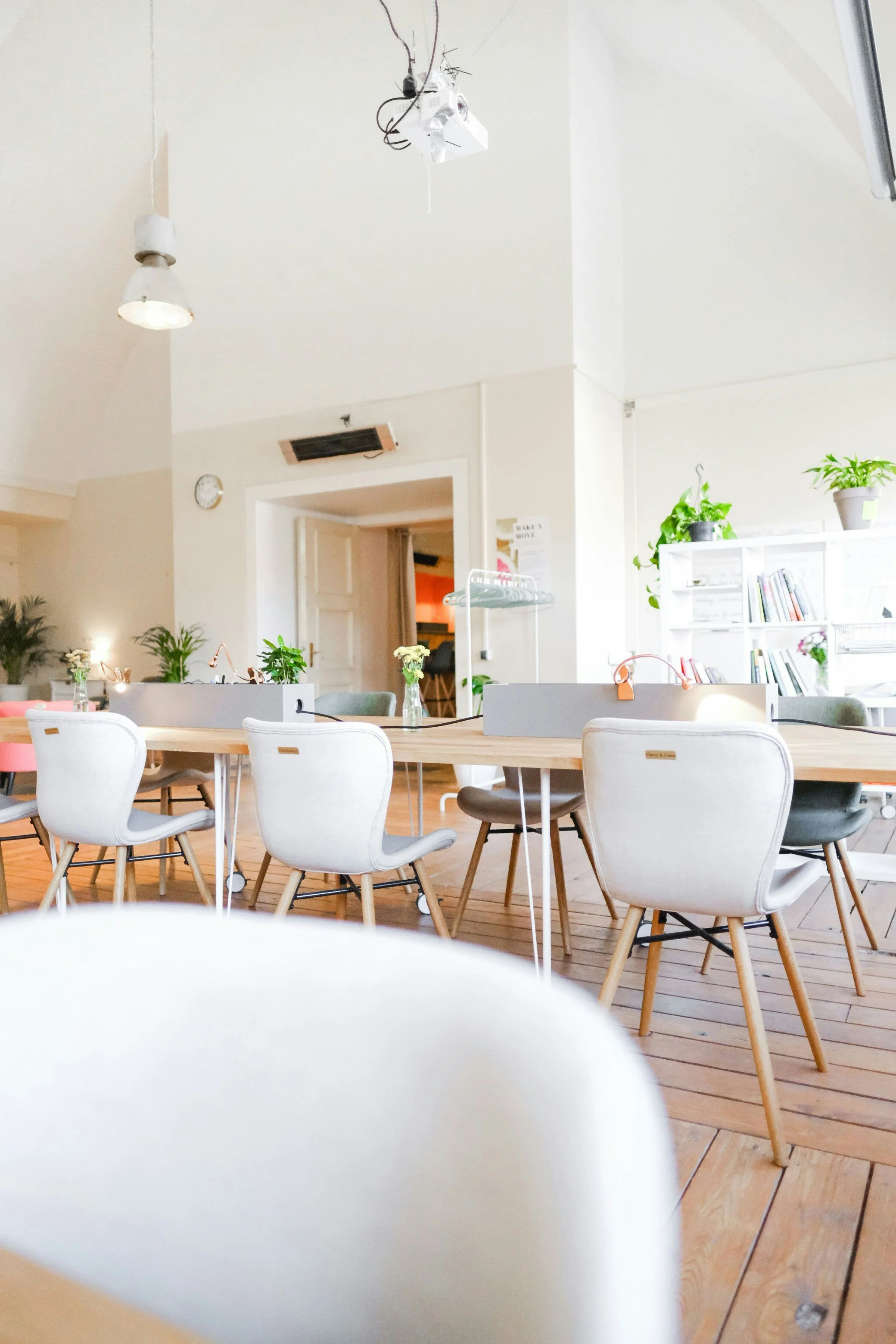 Bright, modern coworking space with white and pastel-colored chairs, wooden tables, and houseplants on shelves and tables.