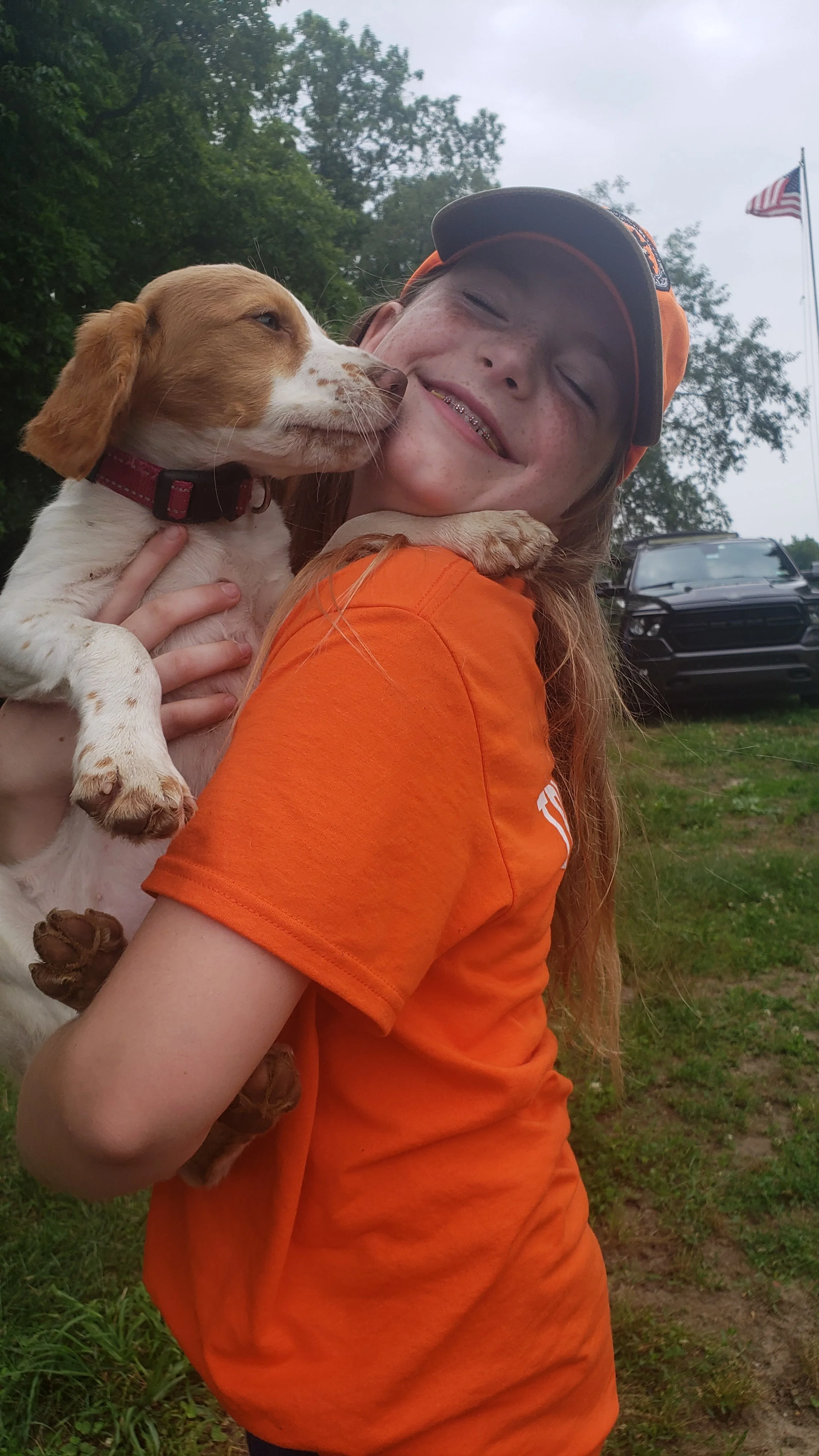 Smiling girl in an orange shirt holding a puppy, outside with trees, a flag, and a truck in the background.