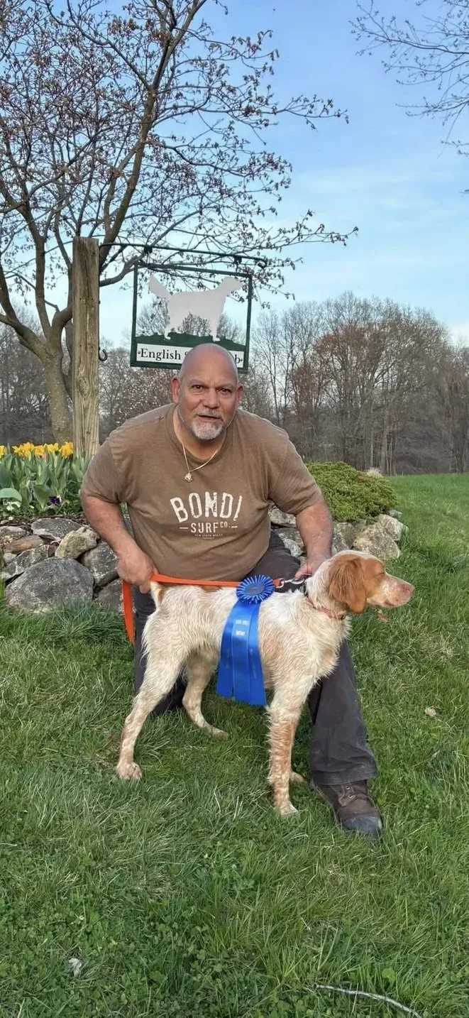 A man kneeling on the grass holding a dog with a blue ribbon. Behind them is a sign with a silhouette of a dog and the words "English" and "sub". The area has trees and flowers.