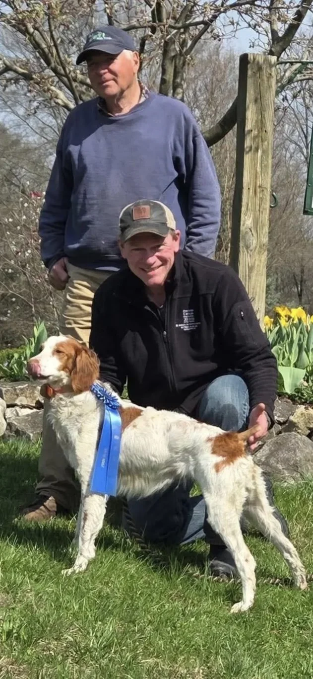 Two men and a dog outdoors in a garden with blooming tulips, a tree, and a wooden post. One man is standing behind, the other squatting next to the dog, which has a blue ribbon around its neck.