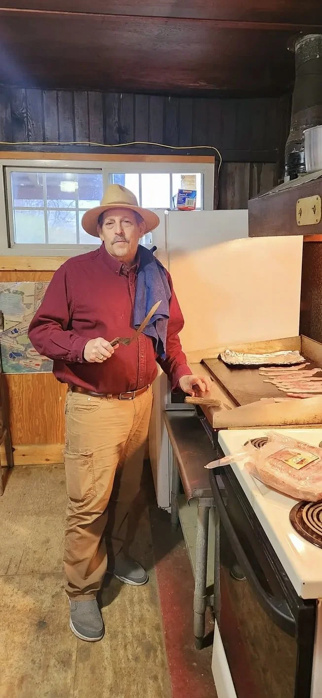A man wearing a wide-brimmed hat and red shirt is standing in a rustic kitchen, cutting raw bacon on a wooden cutting board next to a stove with a package of bacon and a cooked fish. The kitchen has wooden walls and a small window behind him.