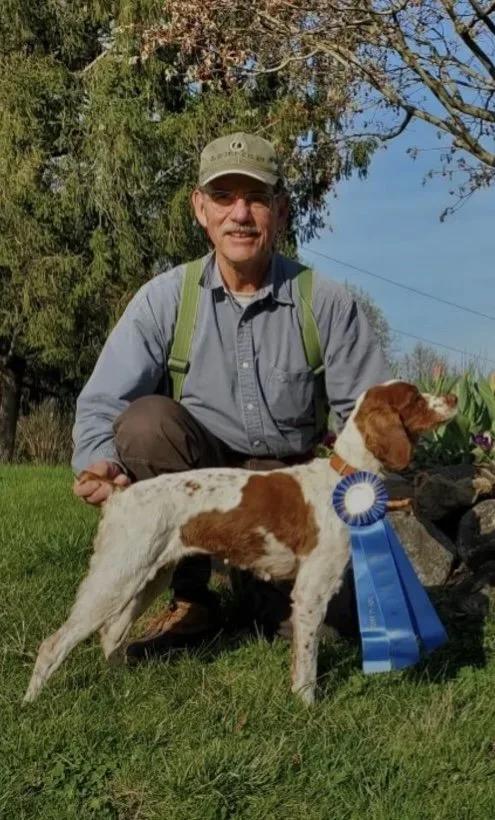 A man kneeling on the grass outdoors, smiling, with a brown and white dog wearing a blue ribbon.