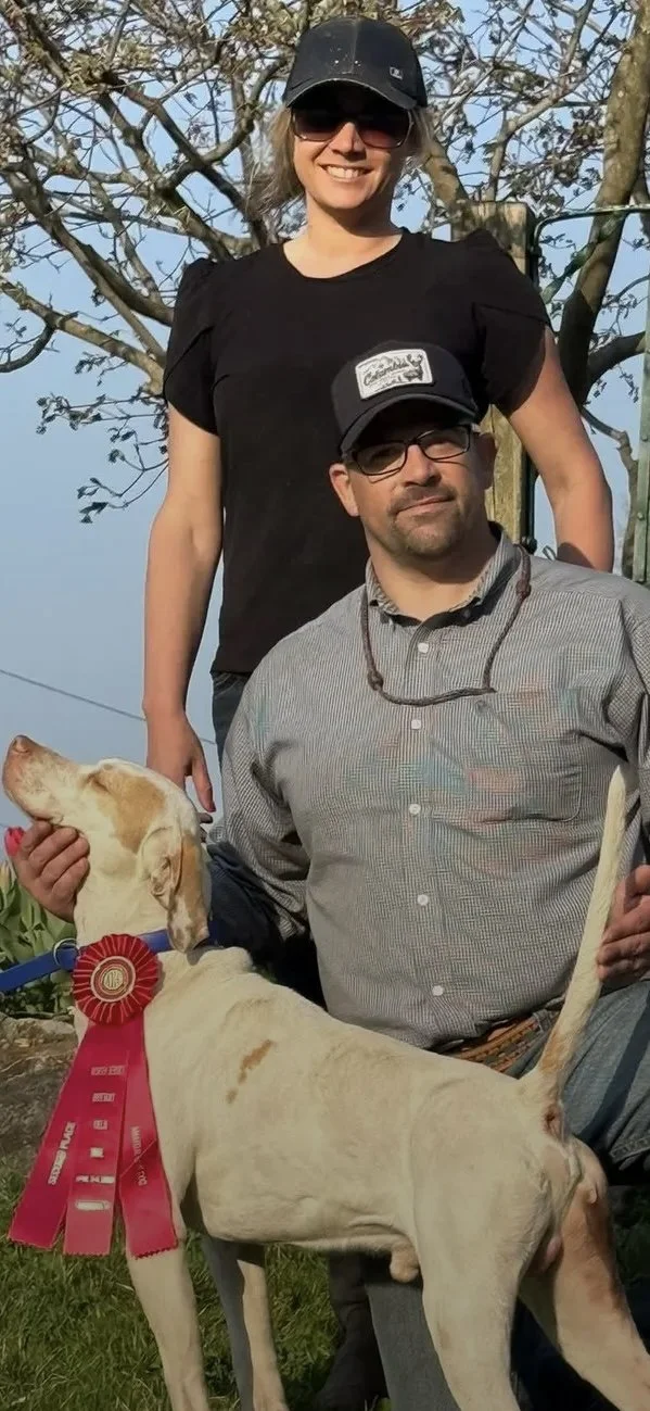 A woman and a man outdoors with a tree in the background. The woman is standing behind the man, wearing sunglasses, a black cap, and a black t-shirt. The man is sitting, wearing glasses, a gray button-up shirt, and a hat. He is holding a yellow hunting dog with a red award ribbon around its neck.