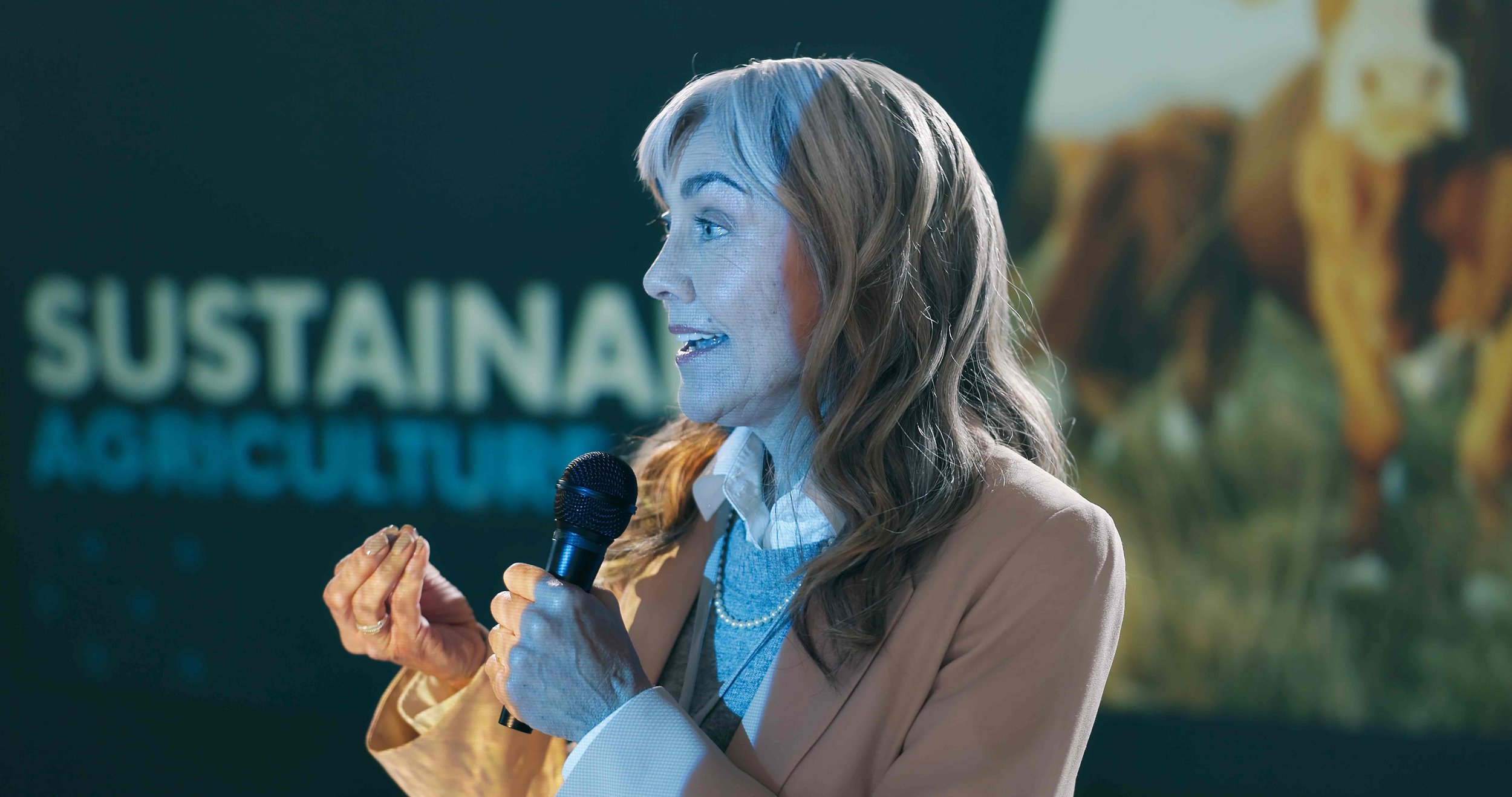 A woman with light brown hair speaking into a microphone at a conference or event, with a blurred background of a display or banner.
