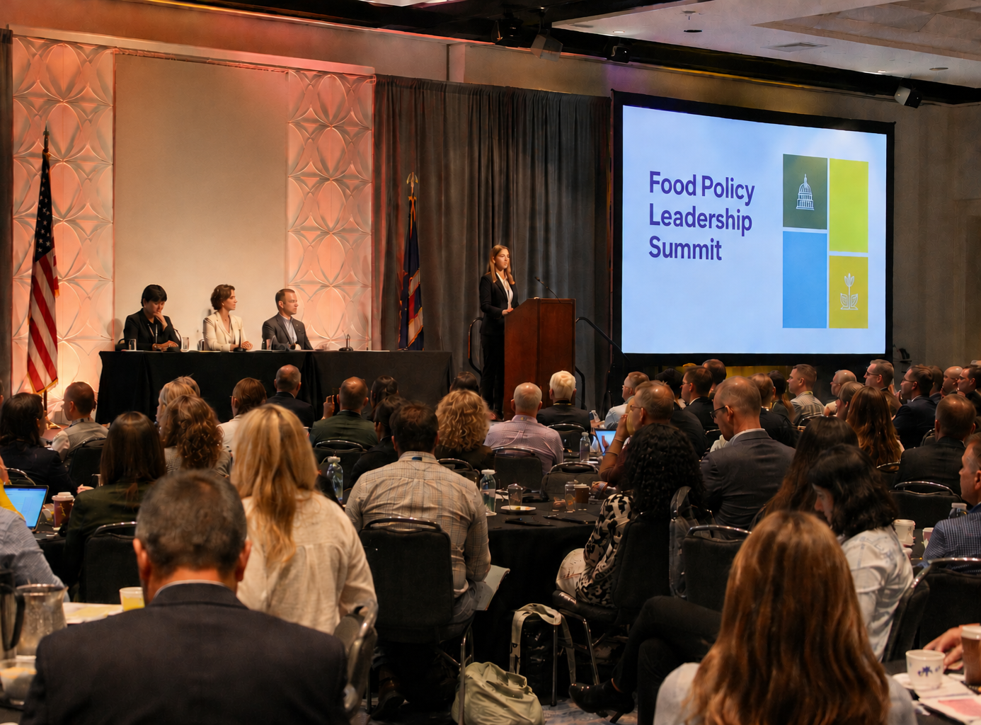 A speaker presenting at the Food Policy Leadership Summit with an audience seated and listening, large screen displaying the event name, and panelists seated at a table on the stage.