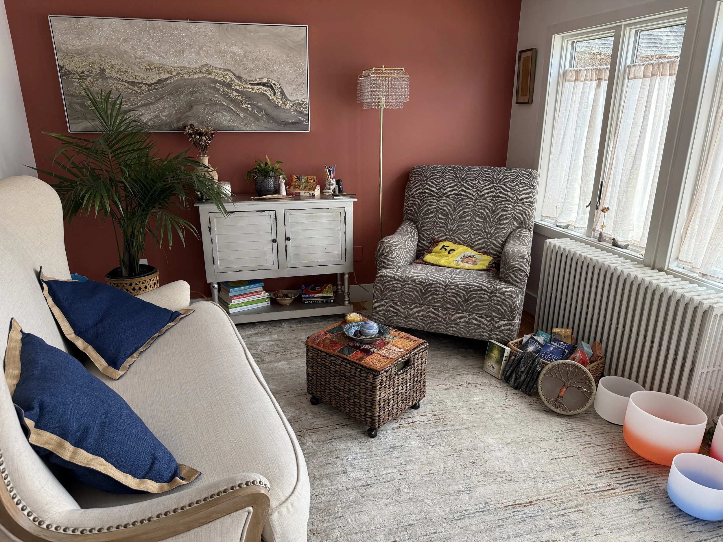 Cozy living room with a beige sofa, blue pillows, a patterned armchair, a white sideboard, a tall floor lamp, and a window with cream curtains. Decorative items include books, plants, colorful bowls, and artwork.