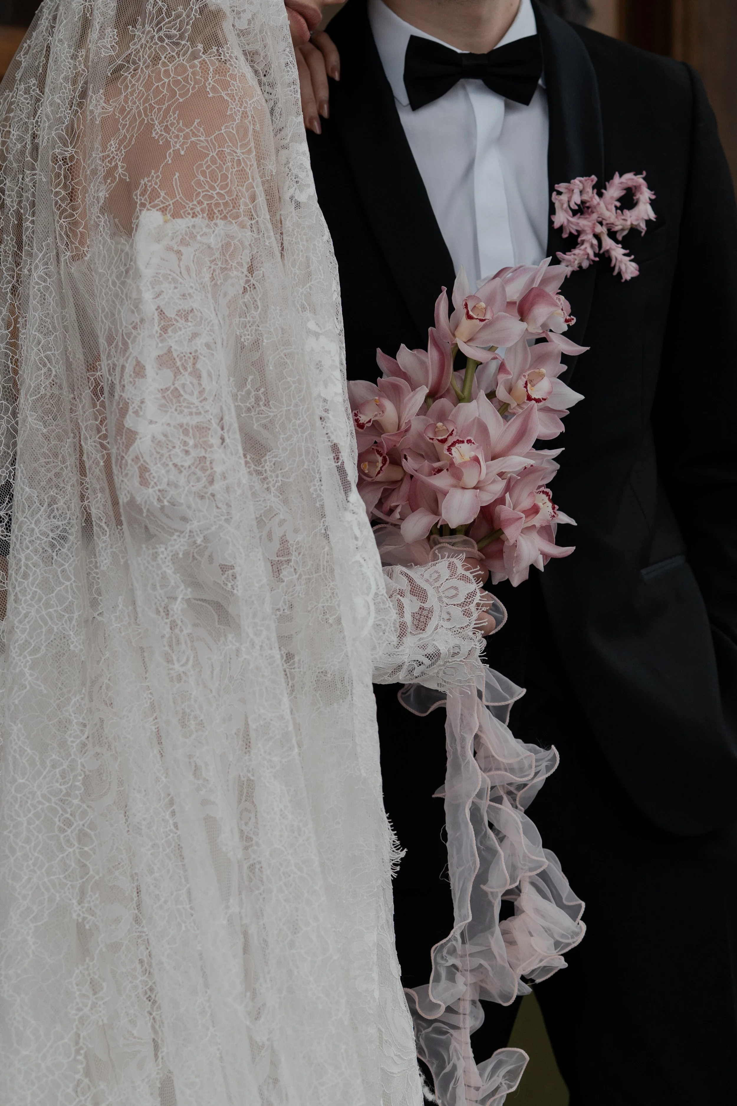 A bride and groom standing close together; the bride is holding a bouquet of pink orchids and the groom is wearing a black tuxedo with a bow tie and pink floral boutonniere.