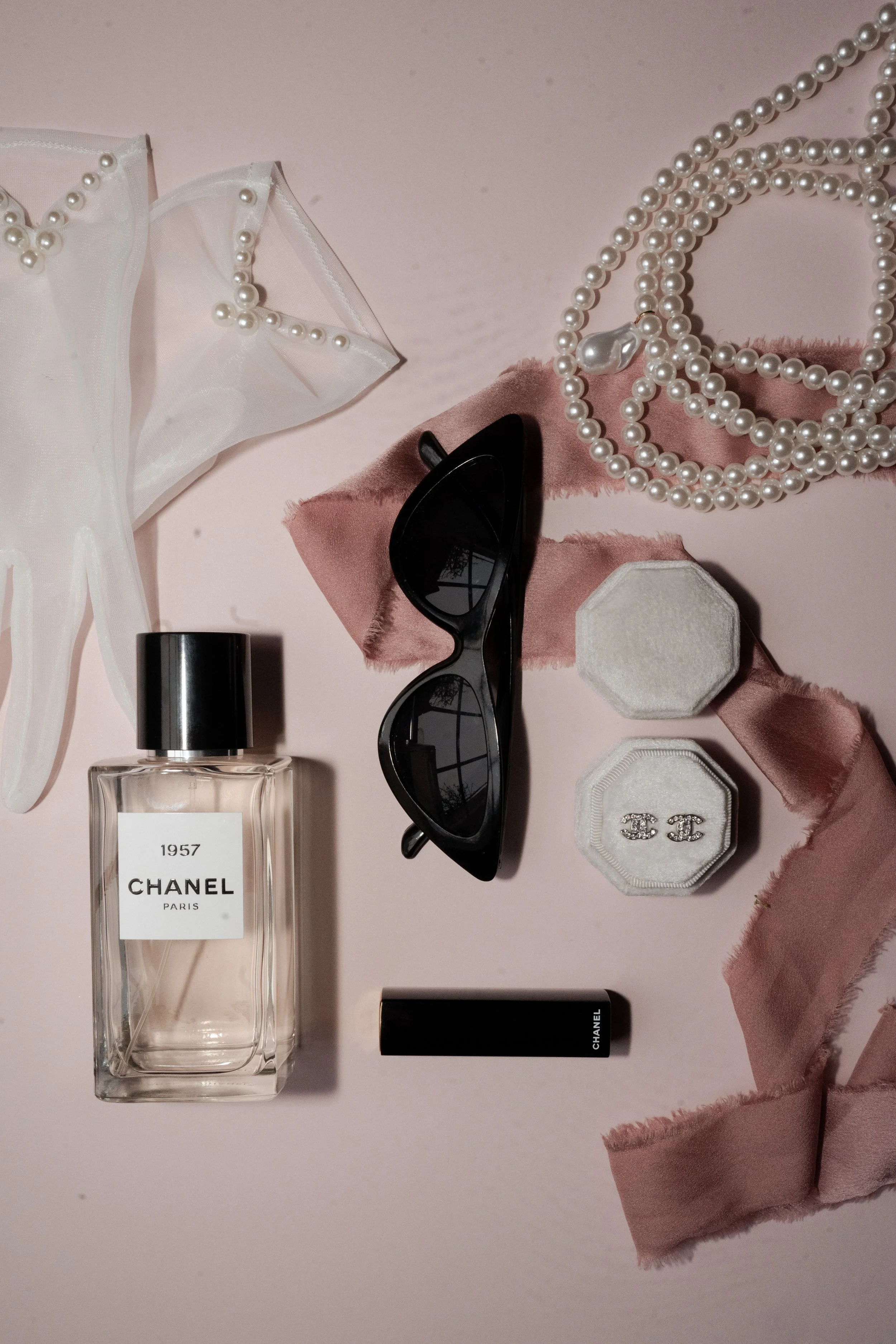 A collection of fashion accessories including a string of pearls, black sunglasses, a bottle of Chanel perfume, a lipstick, a white jewelry box with an 'CC' logo, two pieces of pink fabric, a white fabric with pearl embellishments, and two velvet jewelry boxes on a pink surface.