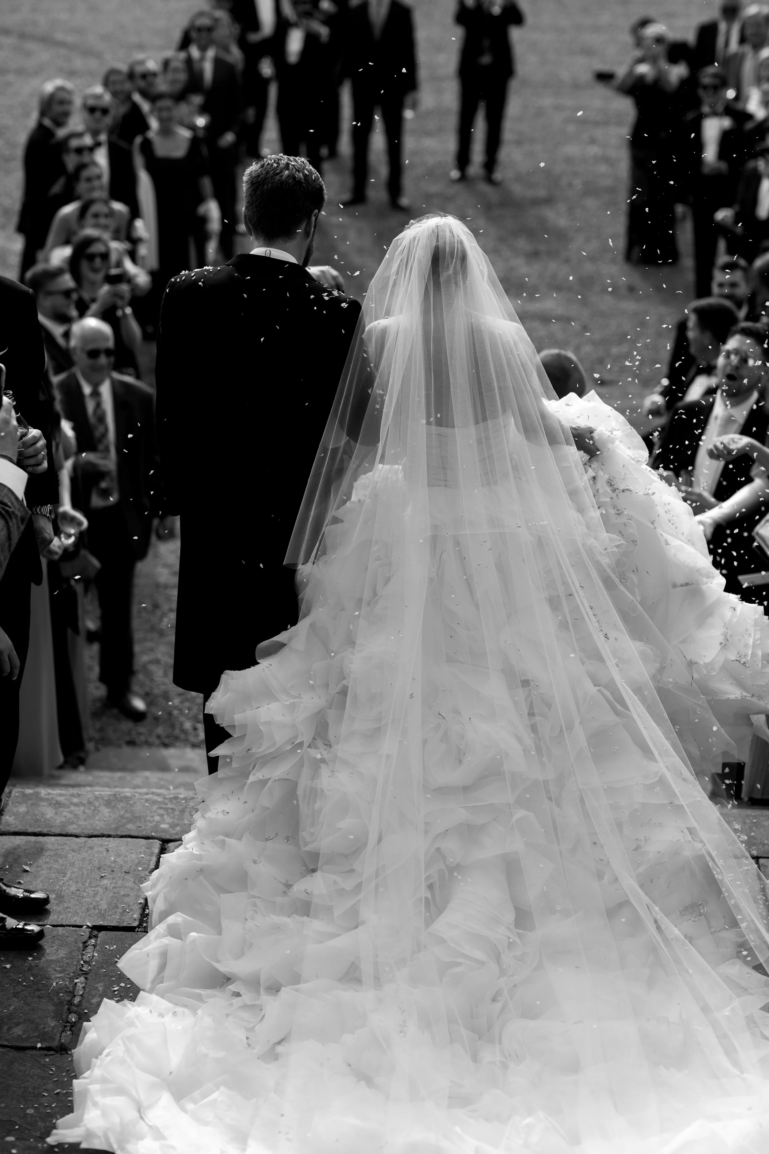 Black and white photo of a bride and groom walking away from the camera on their wedding day, surrounded by guests.