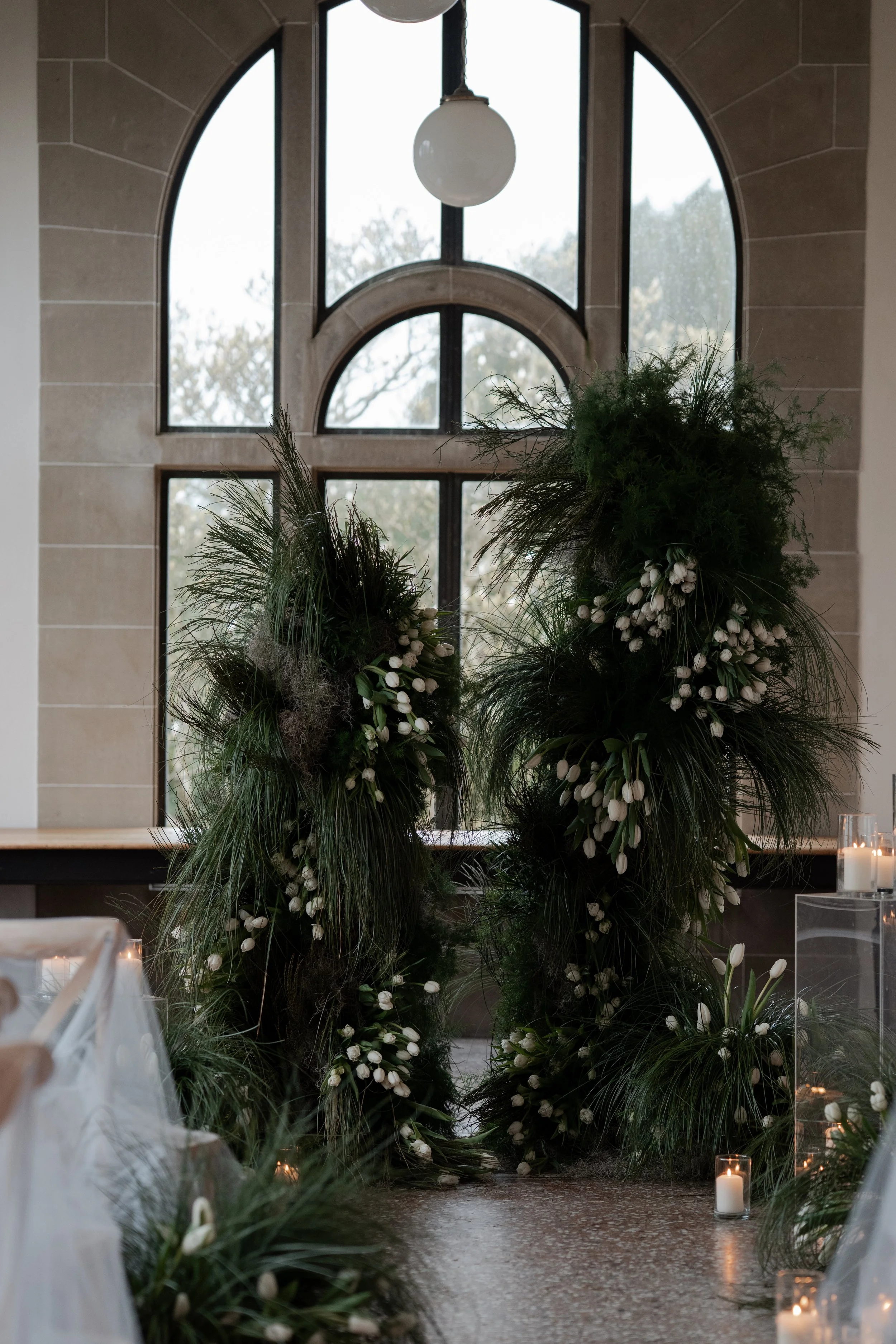 Interior of a building with large arched windows, decorated with tall floral arrangements of white flowers and greenery, and lit candles on the floor.