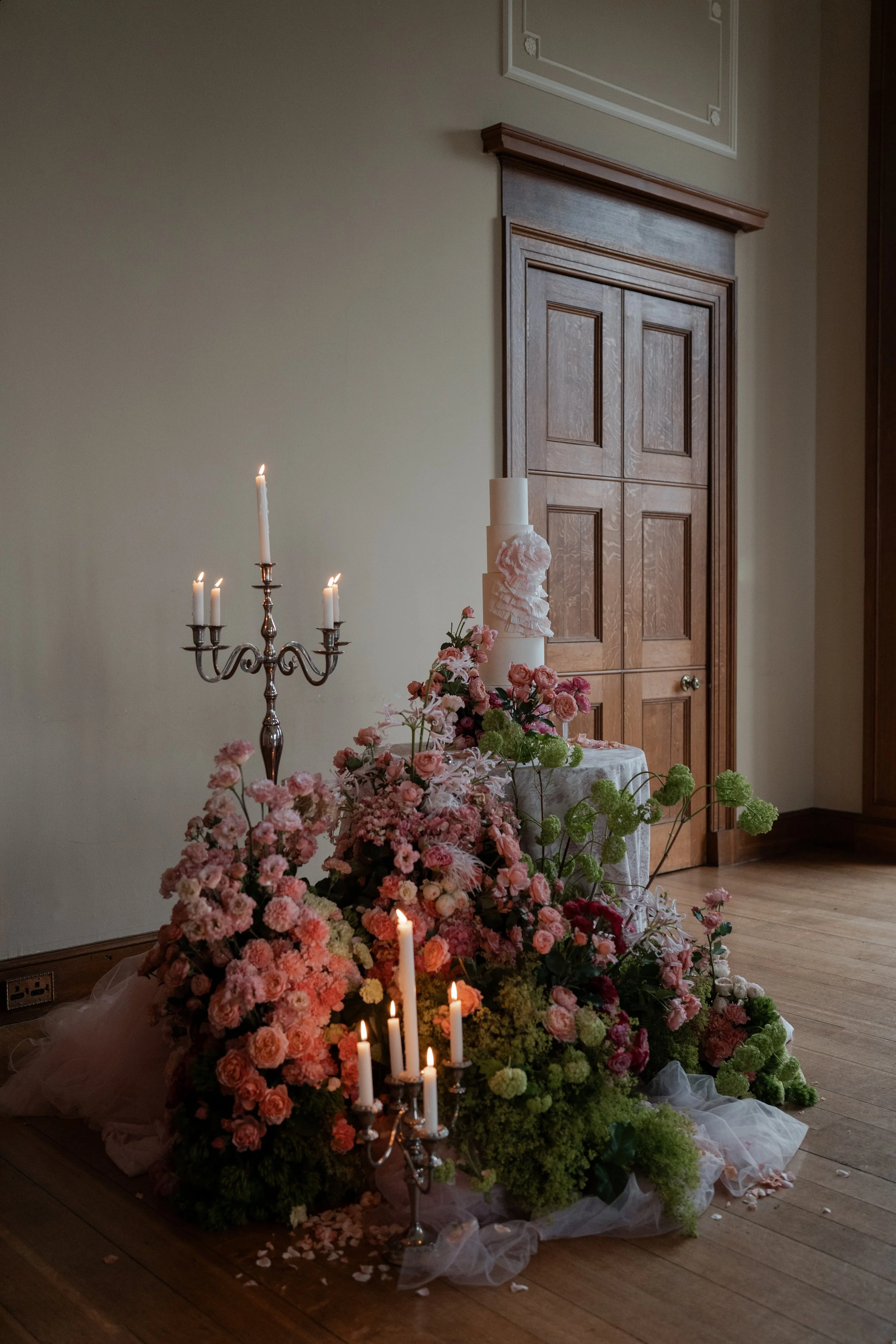A floral arrangement with pink, white, and green flowers, lit candles, and a tall white cake on a stand, set against a wooden door and beige wall in a room.