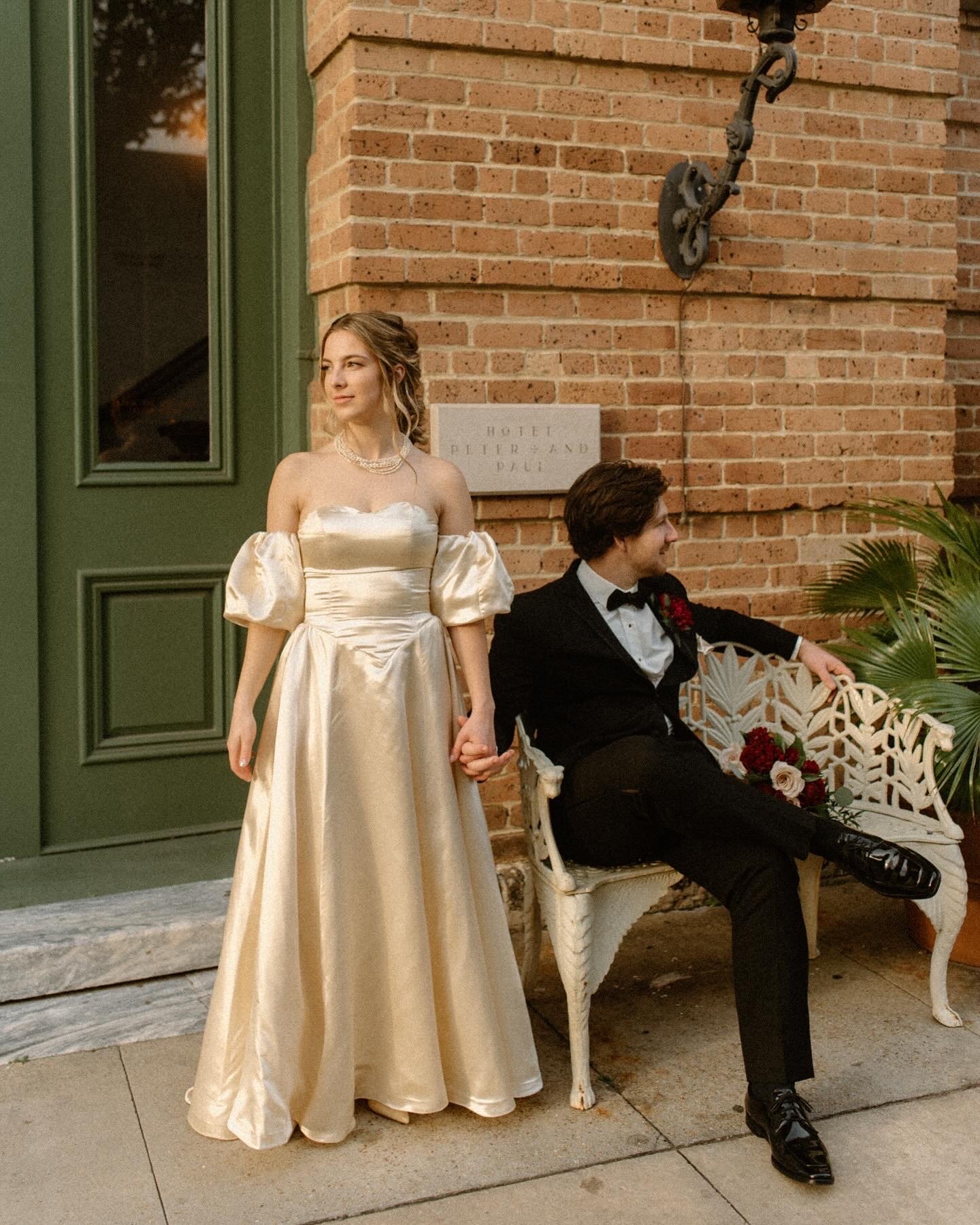 A young woman in a satin off-shoulder gown and pearls holding hands with a young man in a tuxedo sitting on a white vintage bench outside a brick building, with a green door and a potted plant nearby.