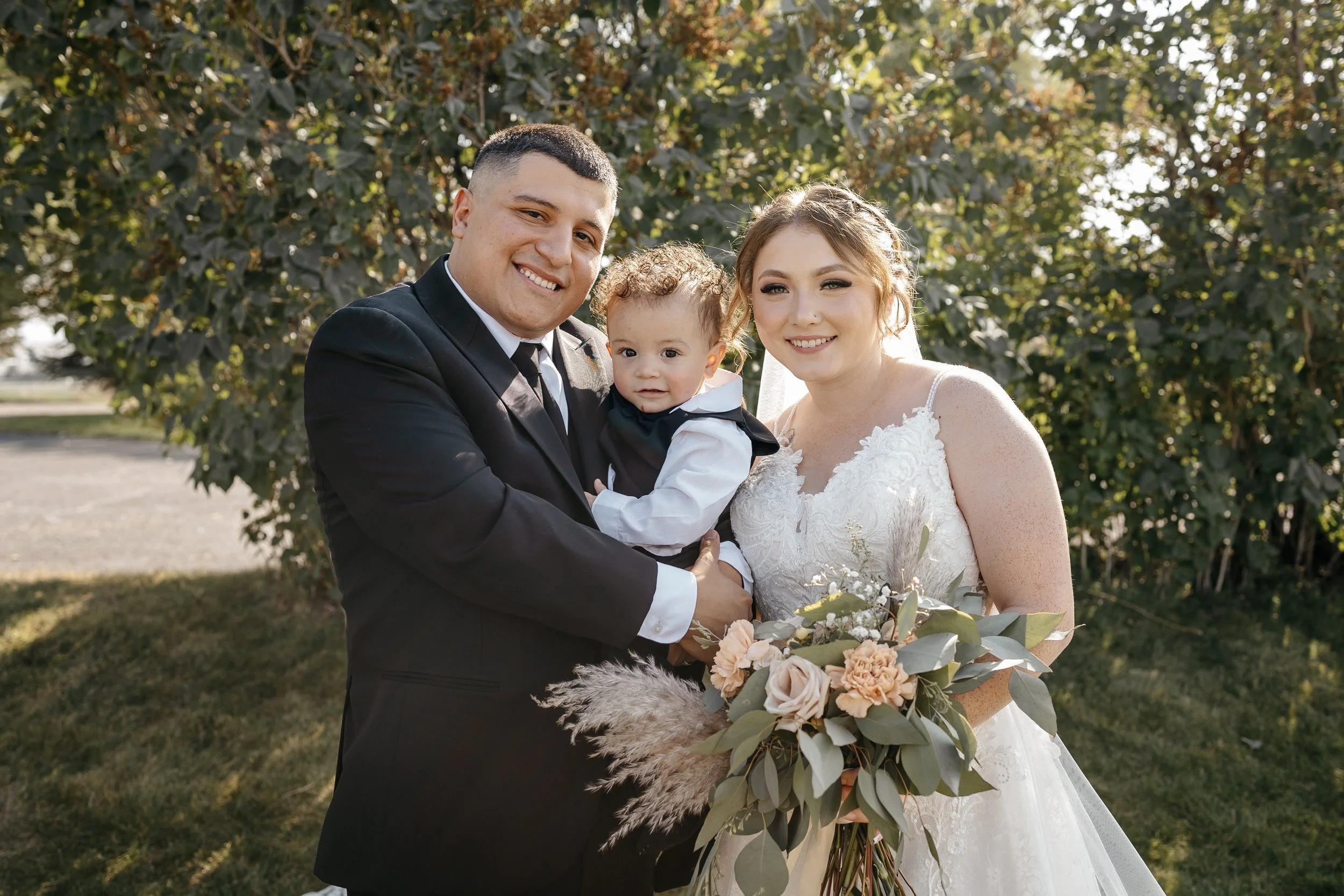 A wedding couple with a young child outdoors, bride in a white wedding dress holding a bouquet, groom in a black suit, and a child in a vest and white shirt, all smiling in front of trees.