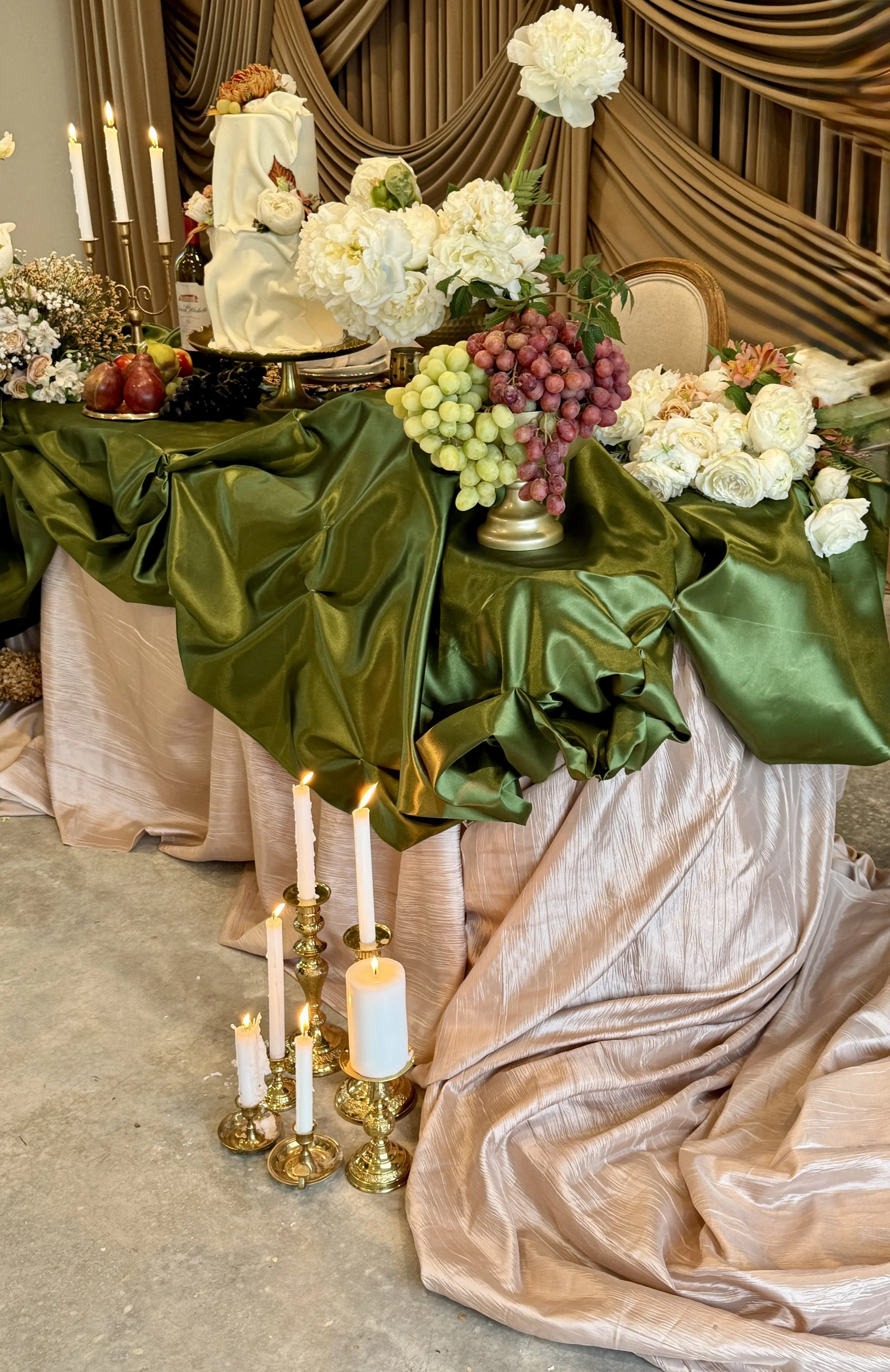 Decorative table with green and pink fabric, flowers, candles, and a cake in the background.