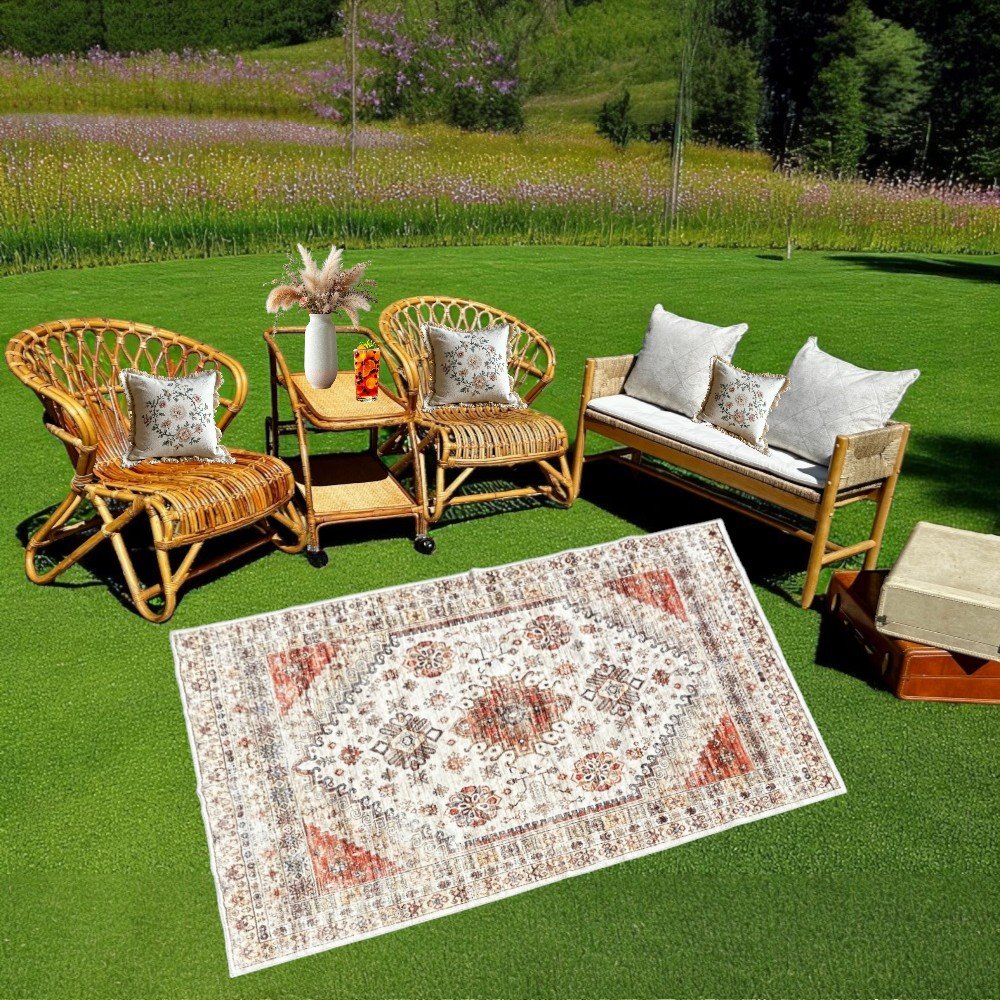 Outdoor seating area with rattan chairs and sofa, decorative pillows, a small table with a vase, and a patterned rug on grass in a garden setting.