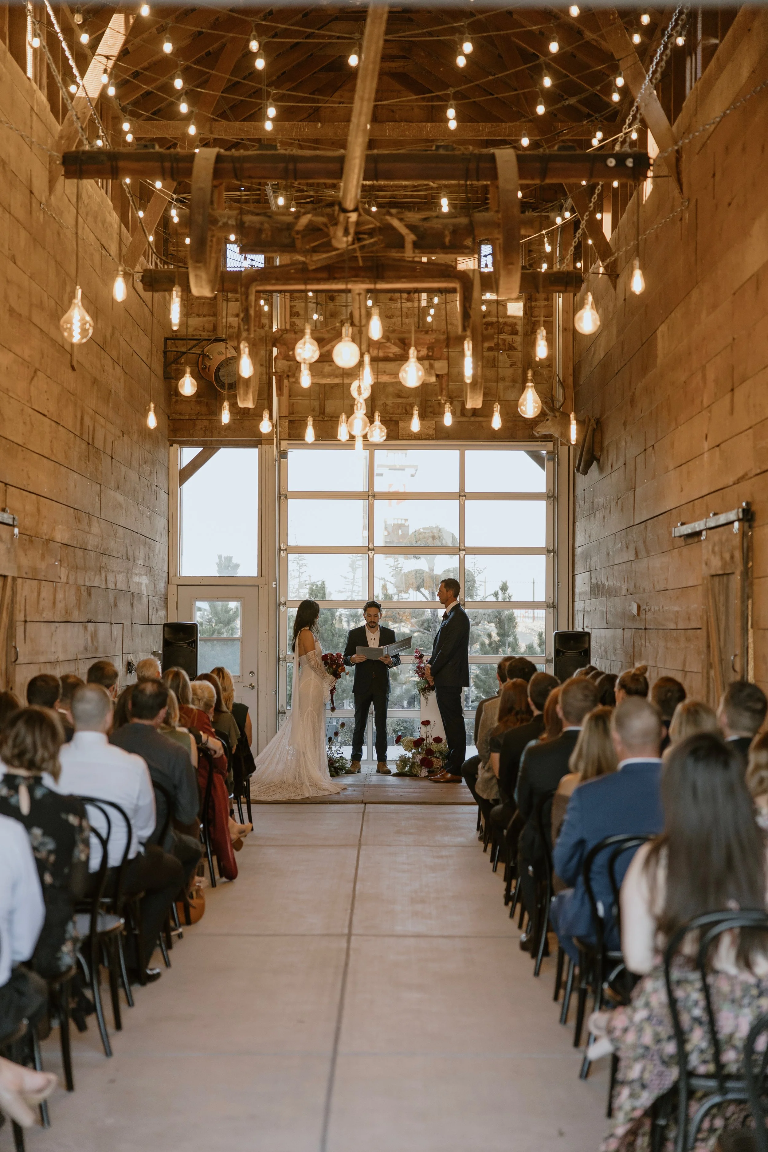 A wedding ceremony taking place inside a rustic barn with wooden walls and string lights hanging from the ceiling. The bride and groom stand at the altar with an officiant, and guests are seated on either side watching the ceremony.