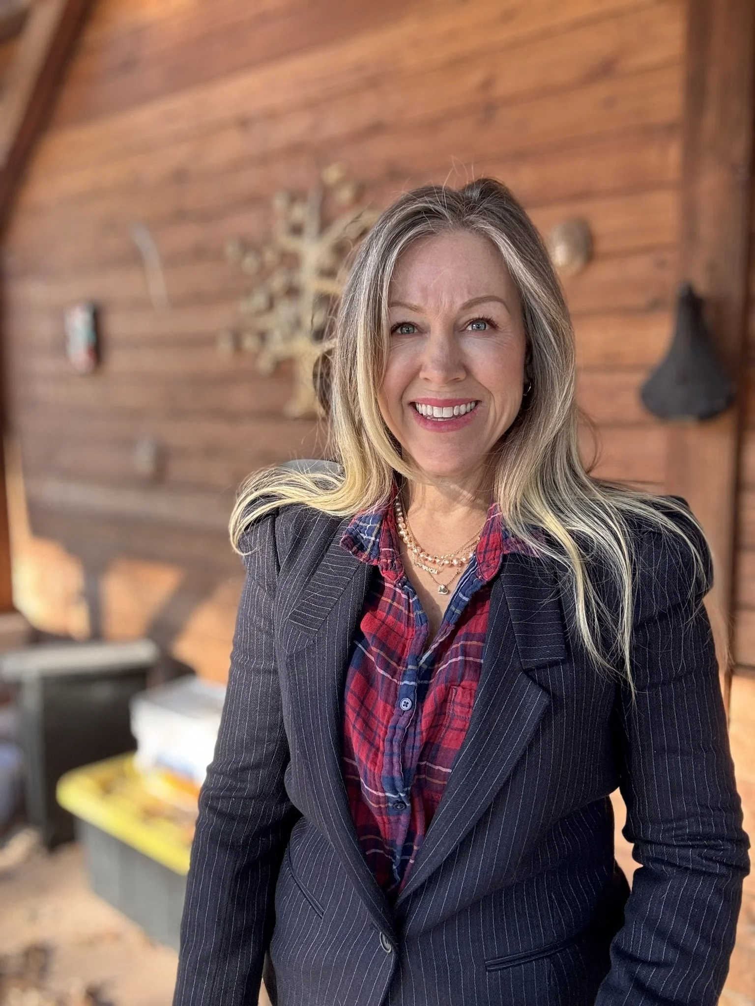 A woman with long blonde hair smiling, wearing a dark pinstripe blazer and a red plaid shirt, standing in front of a wooden wall with wall decorations in the background.