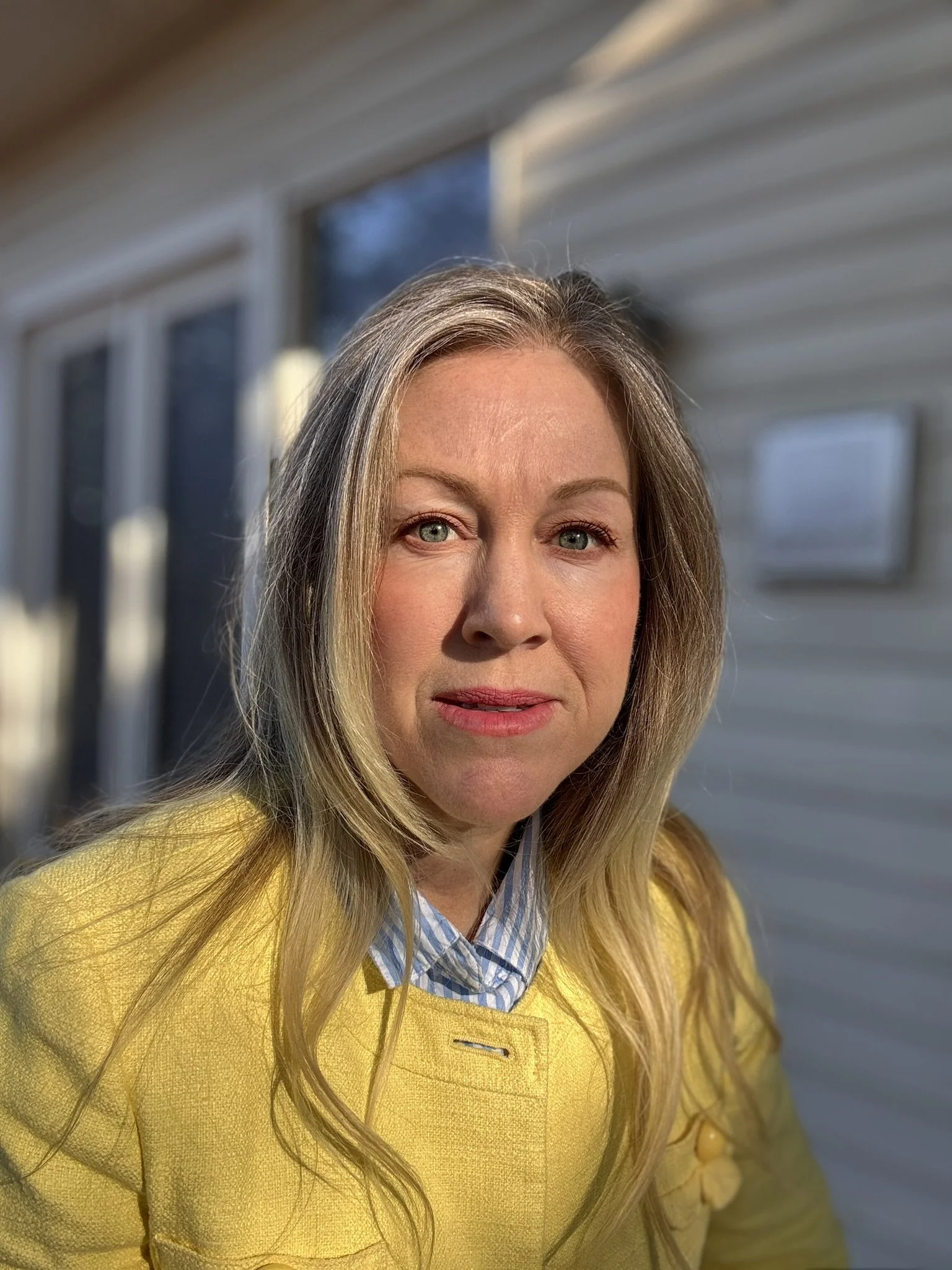 Close-up of a woman with blonde hair and blue eyes wearing a yellow jacket and a striped blue shirt, standing outdoors near a house with horizontal siding, illuminated by sunlight.