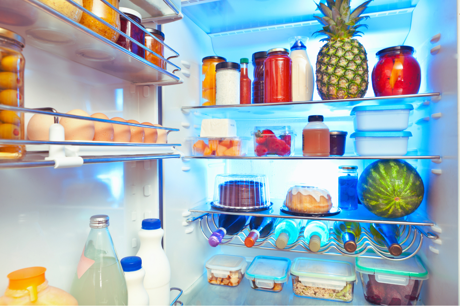 Inside a refrigerator door with various food and drink items including eggs, jars of fruit preserves, bottles of water and juice, a pineapple, a watermelon, yogurt containers, and plastic food storage containers.