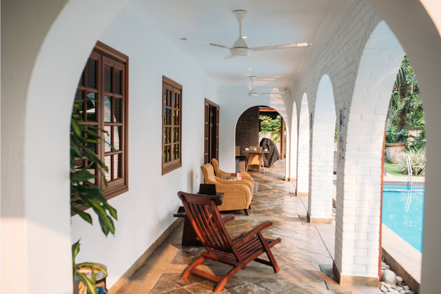A covered outdoor porch with white walls, brown wooden framed windows, and white brick arches. There are wooden and wicker chairs, a table, potted plants, and a swimming pool visible outside.