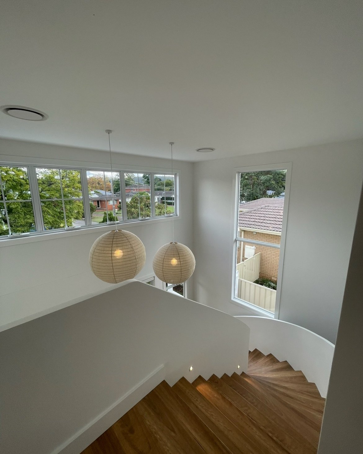 Modern interior staircase with wooden steps, white walls, large windows, and hanging spherical lights.