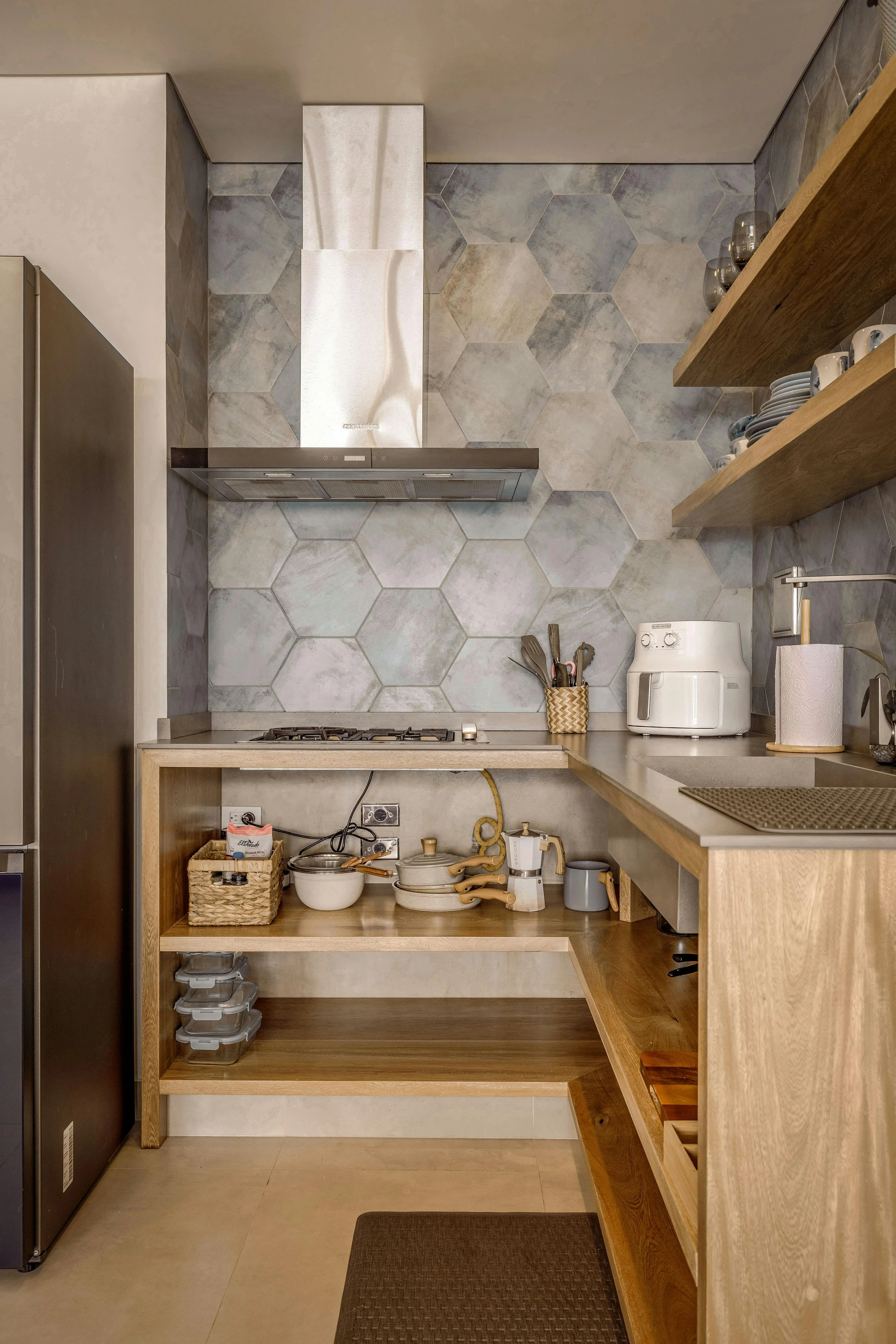 A kitchen with hexagonal wall tiles, wooden shelves, and a stainless steel range hood over a gas stove. The countertops feature a white toaster, a paper towel holder, and various kitchen items, with a wooden open shelf holding bowls, utensils, and containers.