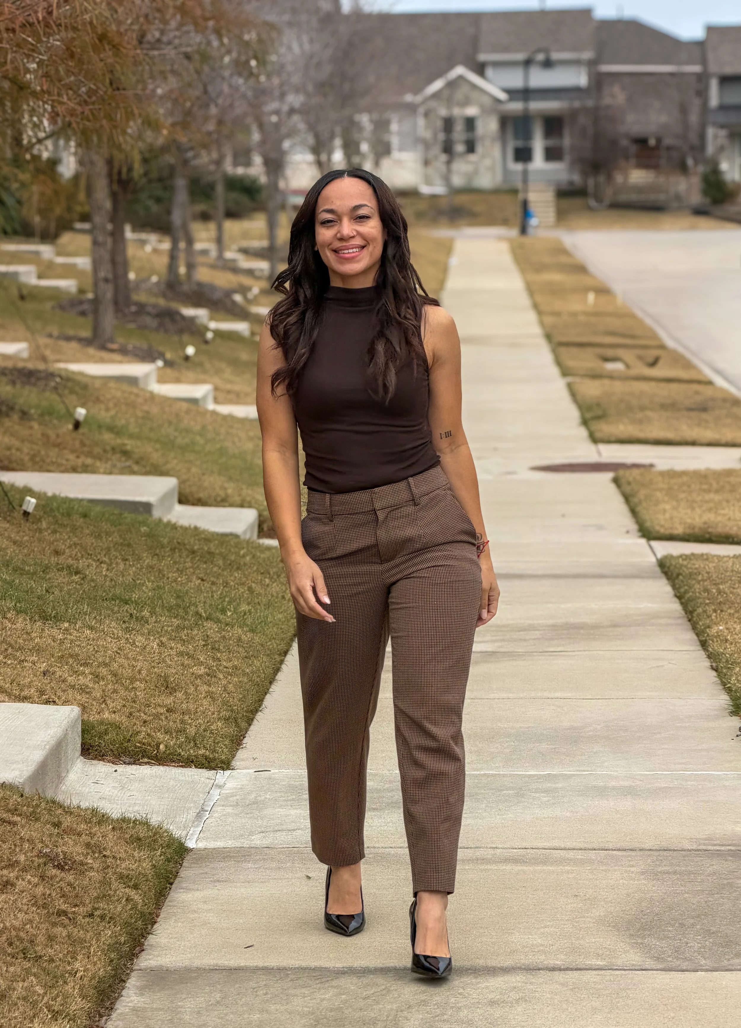 A woman walking on a sidewalk in a residential neighborhood, smiling, with houses and trees in the background.