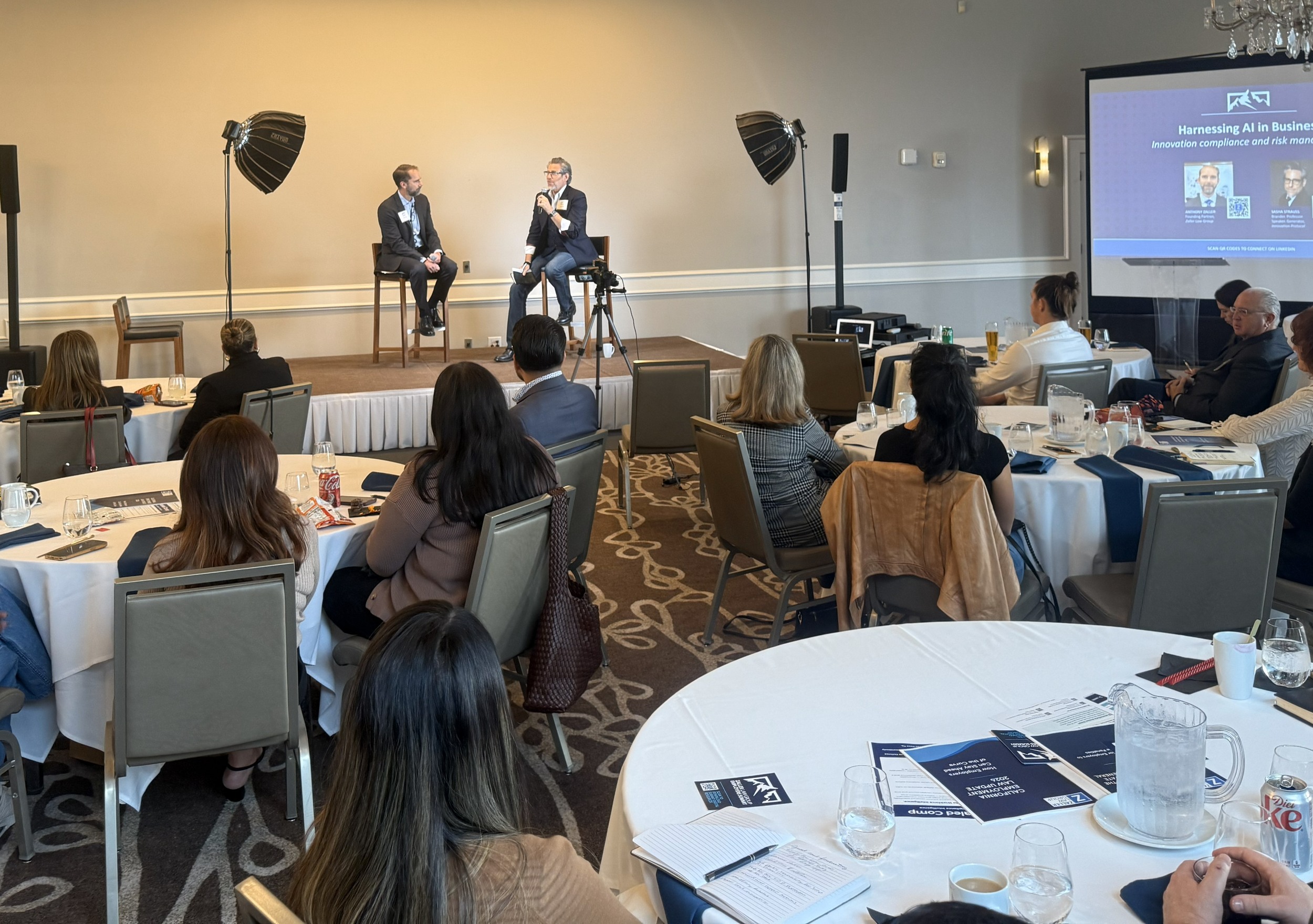 A conference room with a stage where two men are seated and speaking, one with a microphone. Audience members are seated at round tables, and a large screen displays a presentation titled 'Harnessing AI in Business.' The room is well-lit with chandeliers and professional lighting.