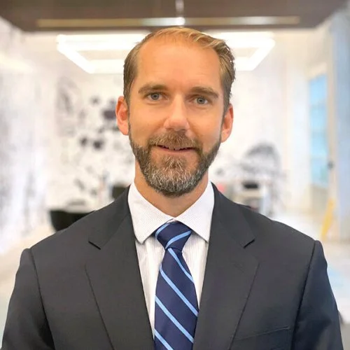 A man in a dark suit and tie standing in an office with modern decor.