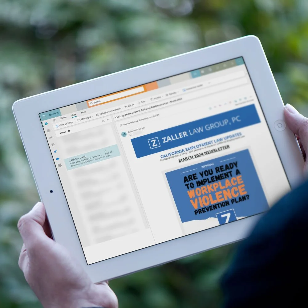 Person holding a white tablet displaying an email newsletter about workplace violence prevention from Zaller Law Group, with a background of blurred outdoor greenery.