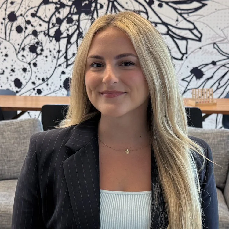 Young woman with long blonde hair, smiling slightly, wearing a dark pinstripe blazer and a white top, sitting in a modern office or lounge area with abstract black and white wall art in the background.