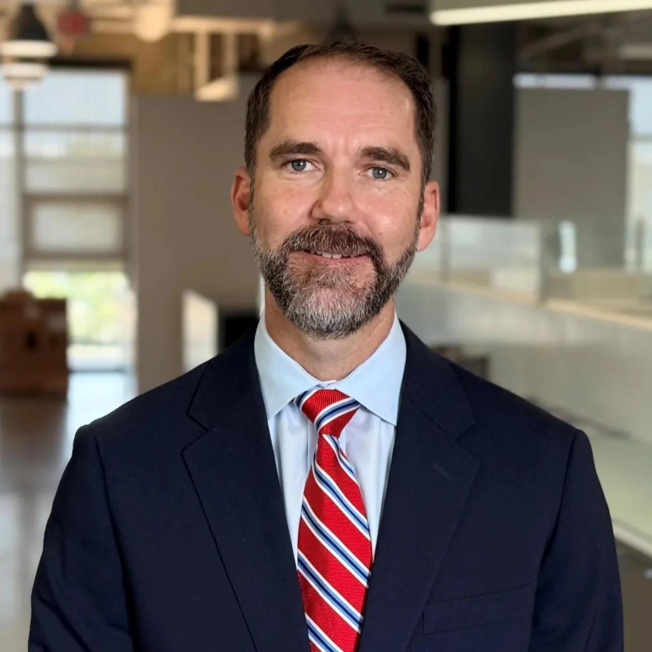 A man with a beard and short light brown hair, wearing a dark suit, white shirt, and striped blue tie, standing in a modern office with white walls and artwork, smiling at the camera.