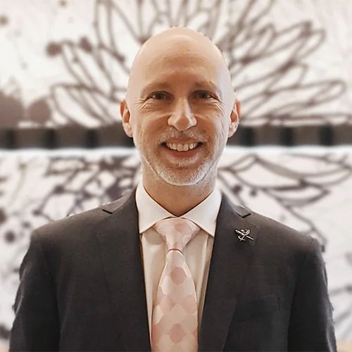A smiling man in a suit and tie standing in front of a blurred background with a large, abstract floral pattern.