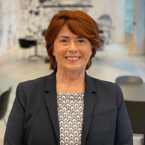 A smiling woman with short red hair wearing a dark blazer and patterned blouse, standing in a modern office setting.