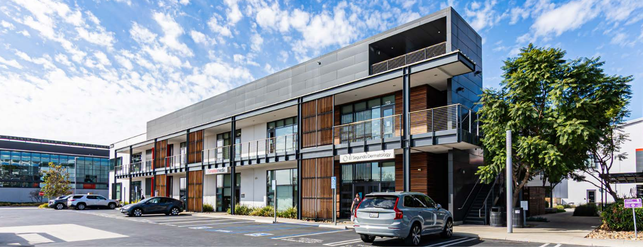 Modern multi-story commercial building with parking lot and cars, blue sky, trees, and people walking.