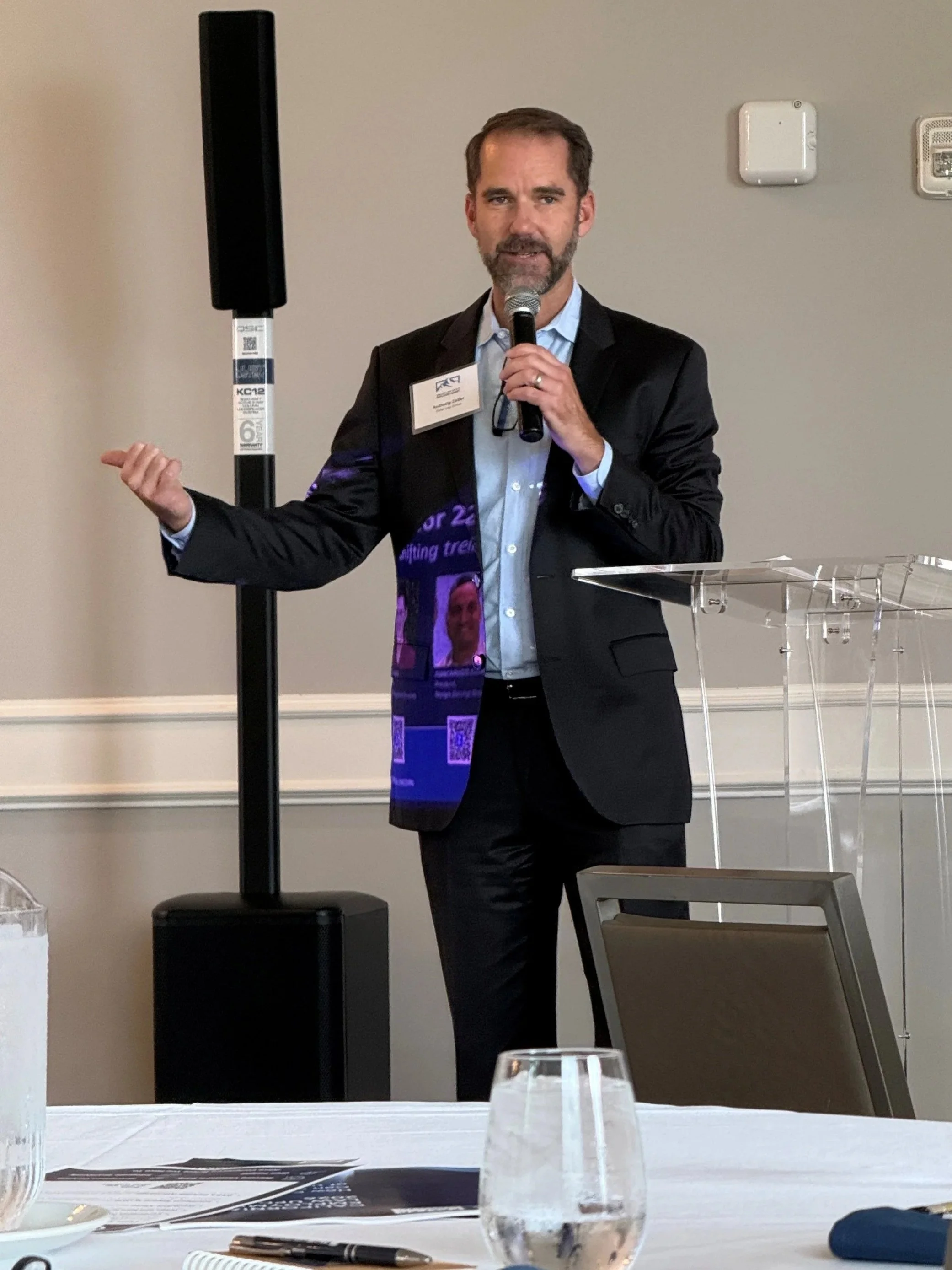 A man in a suit with a name tag is giving a presentation, speaking into a microphone, standing behind a clear podium in a conference room.