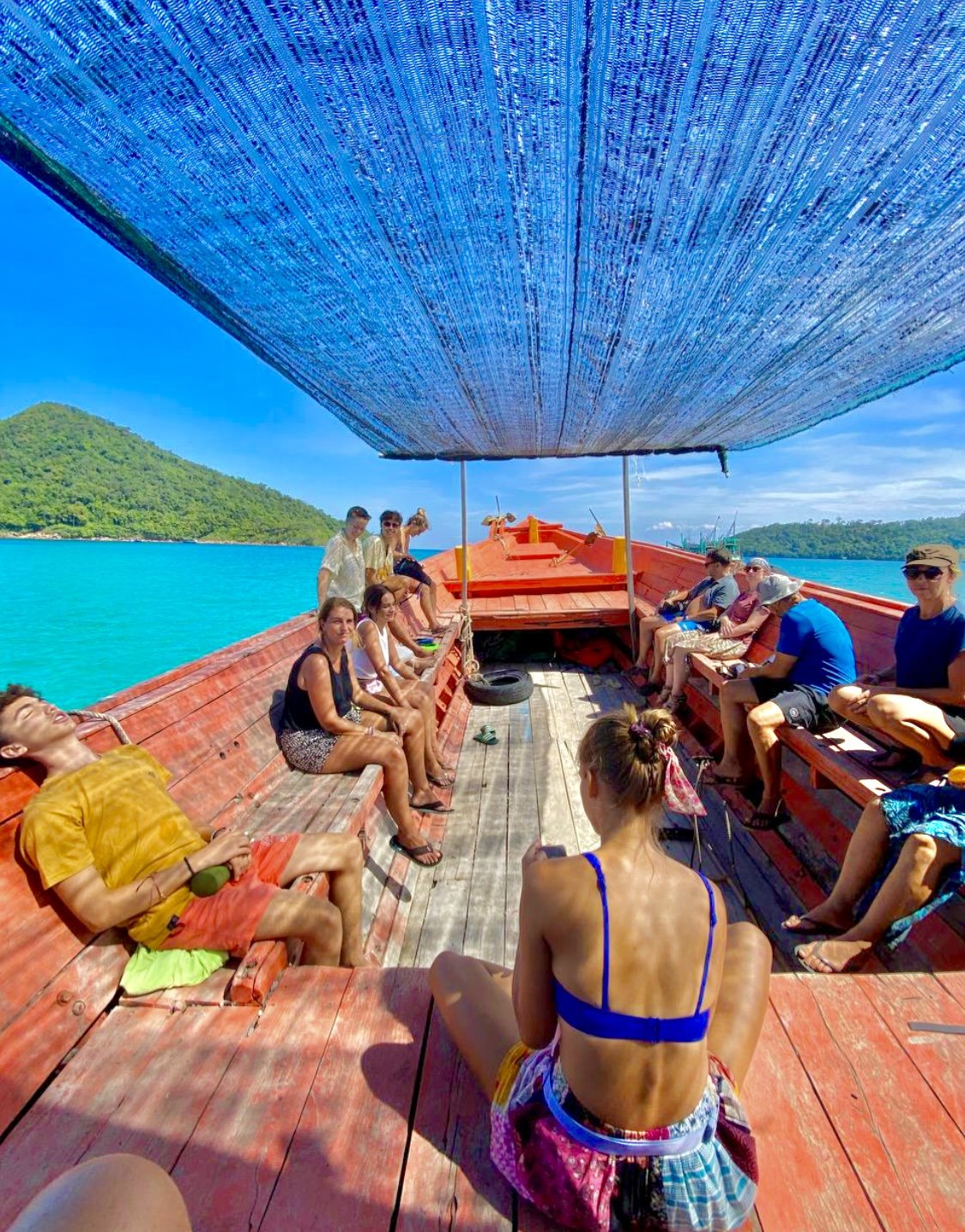 Group of people sitting on a boat with a blue canopy, surrounded by turquoise water and an island in the background.