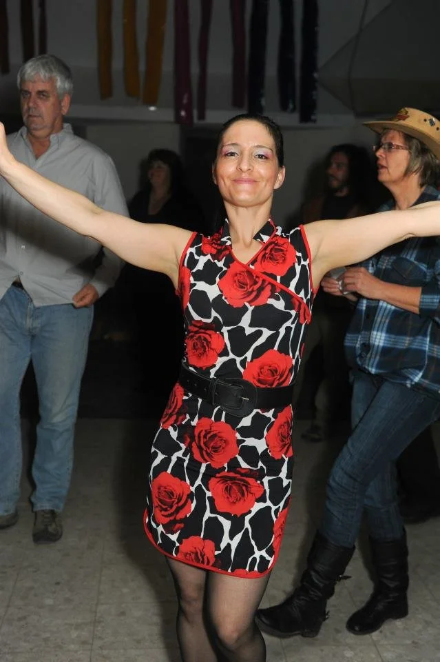 A woman in a sleeveless dress with a red rose pattern poses with her arms outstretched at a social gathering, with other attendees visible in the background.