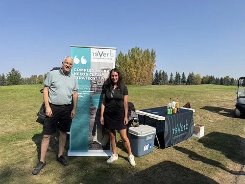Two people standing outdoors next to a promotional banner for 9Verb, with a table of drinks and snacks nearby, in a grassy field with trees and a vehicle in the background.