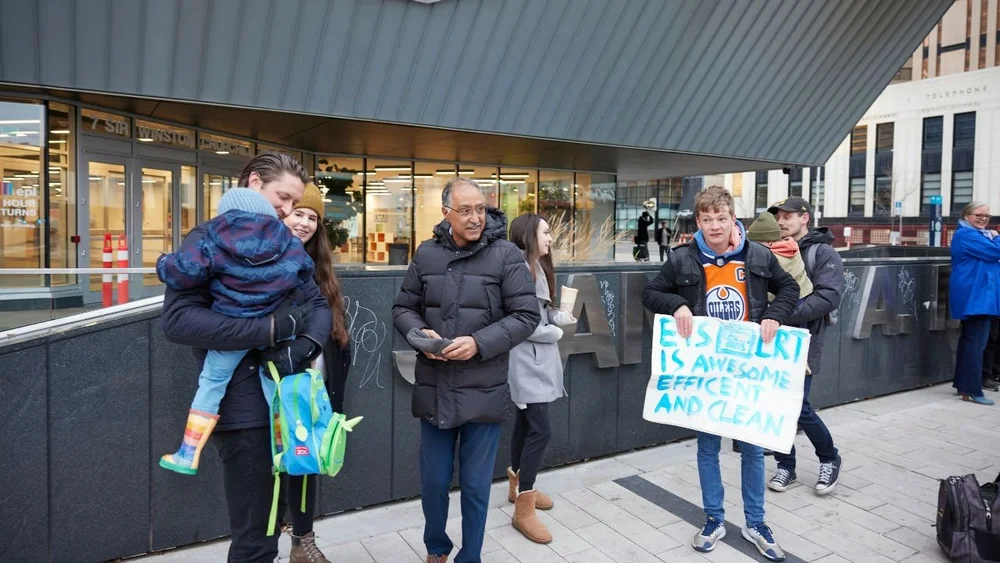 Riders waiting at a Valley Line Southeast station, one holding a hand-made sign